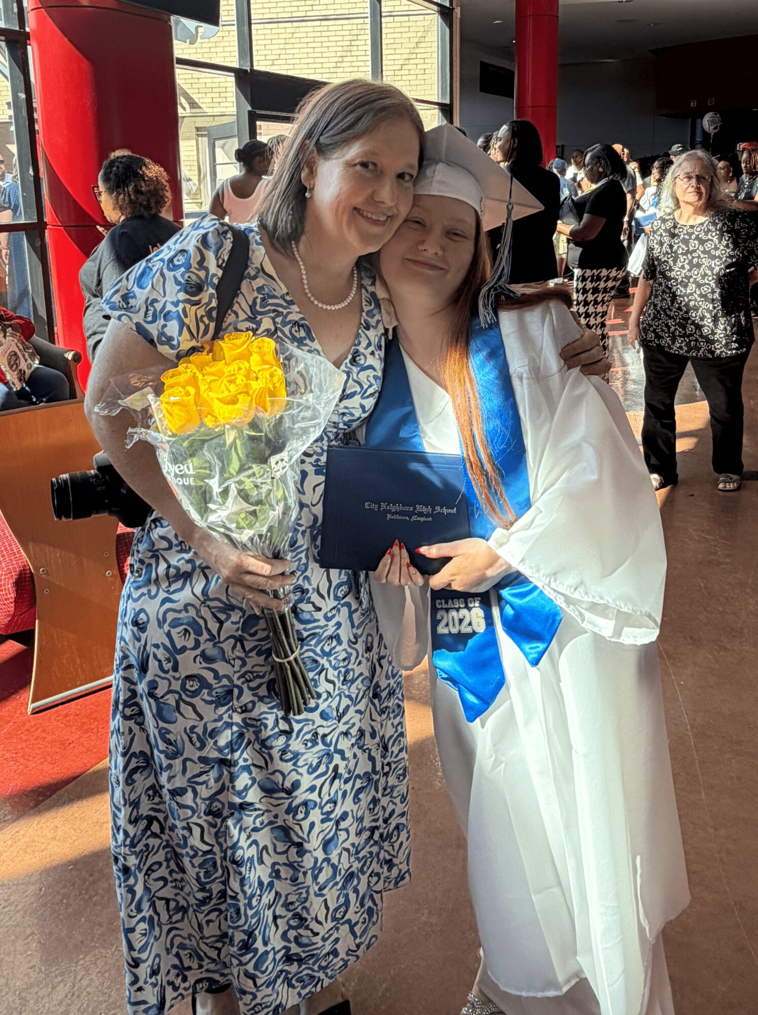 Two women, one in a graduation gown and cap, and the other in a patterned dress, smiling and holding flowers, in a busy indoor setting.