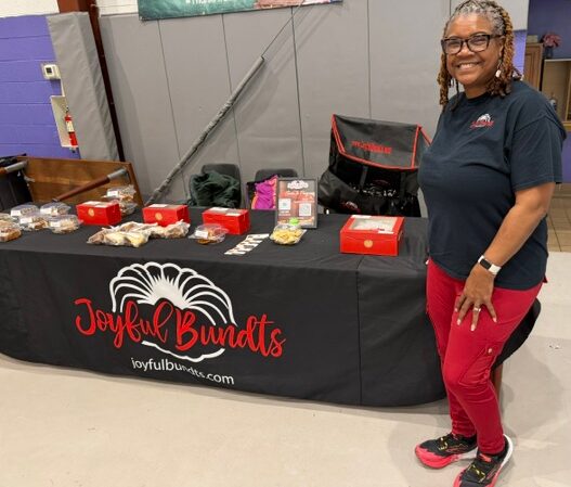 Woman standing next to a table with baked goods and a Joyful Bundts banner, smiling, in an indoor setting.