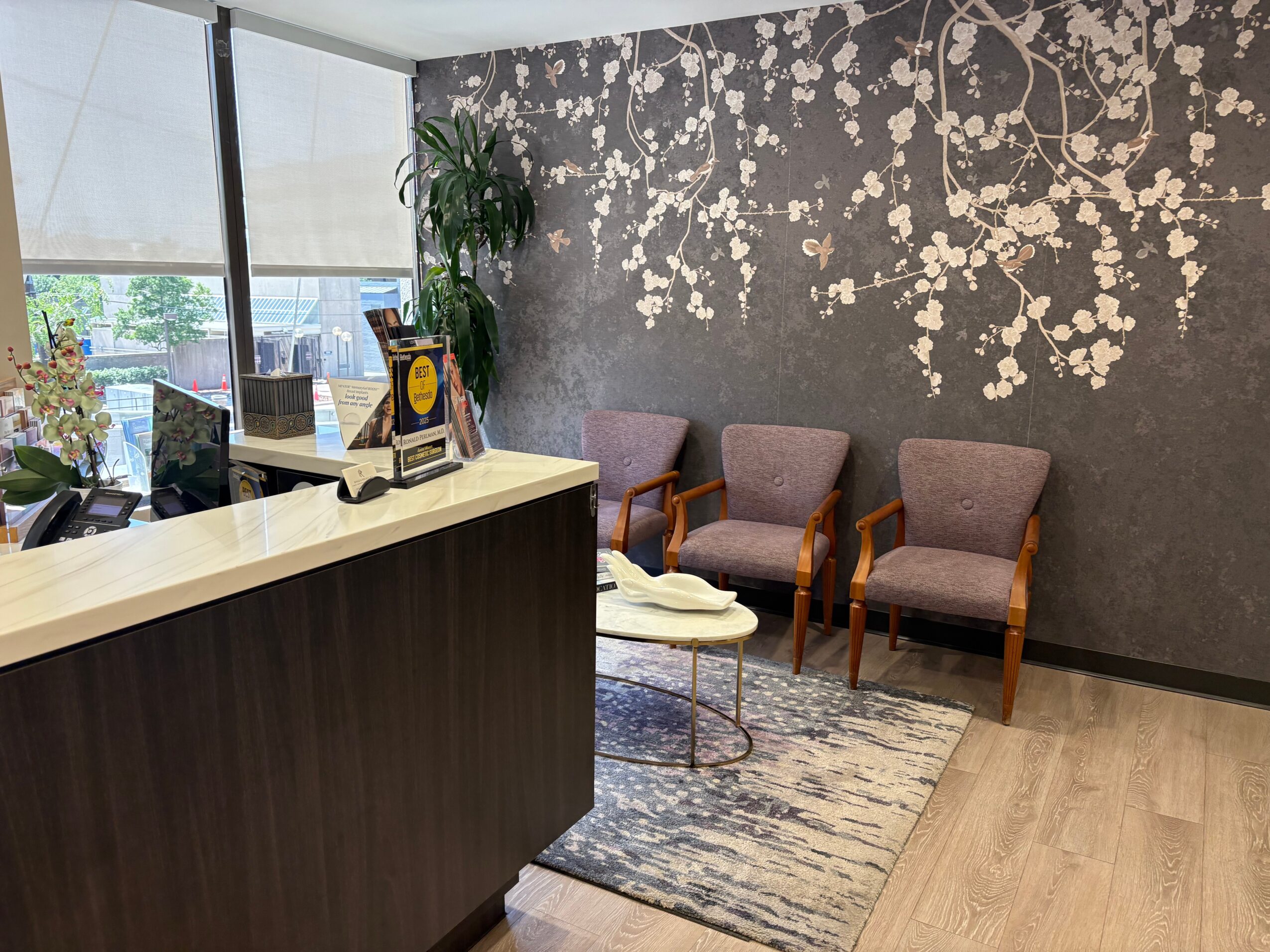 Interior of a waiting area with three chairs, a small table, a rug, and a wall with floral wallpaper.