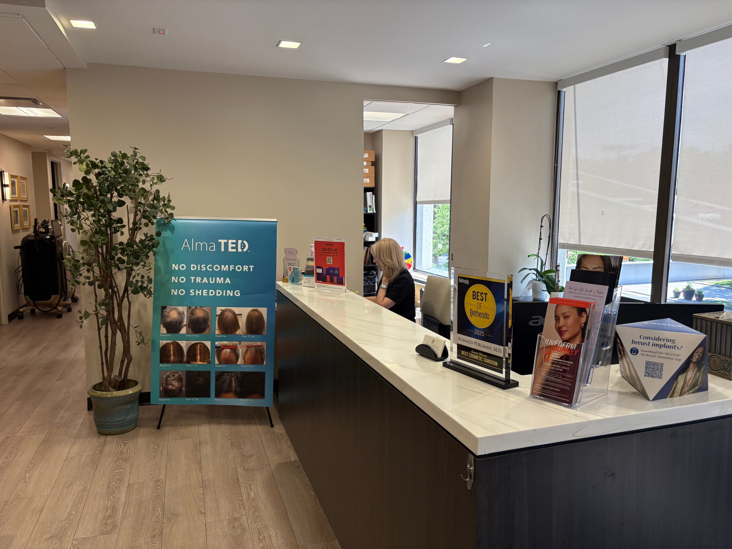 Reception area with a long white counter, a person sitting behind it, and large windows with a view outside.