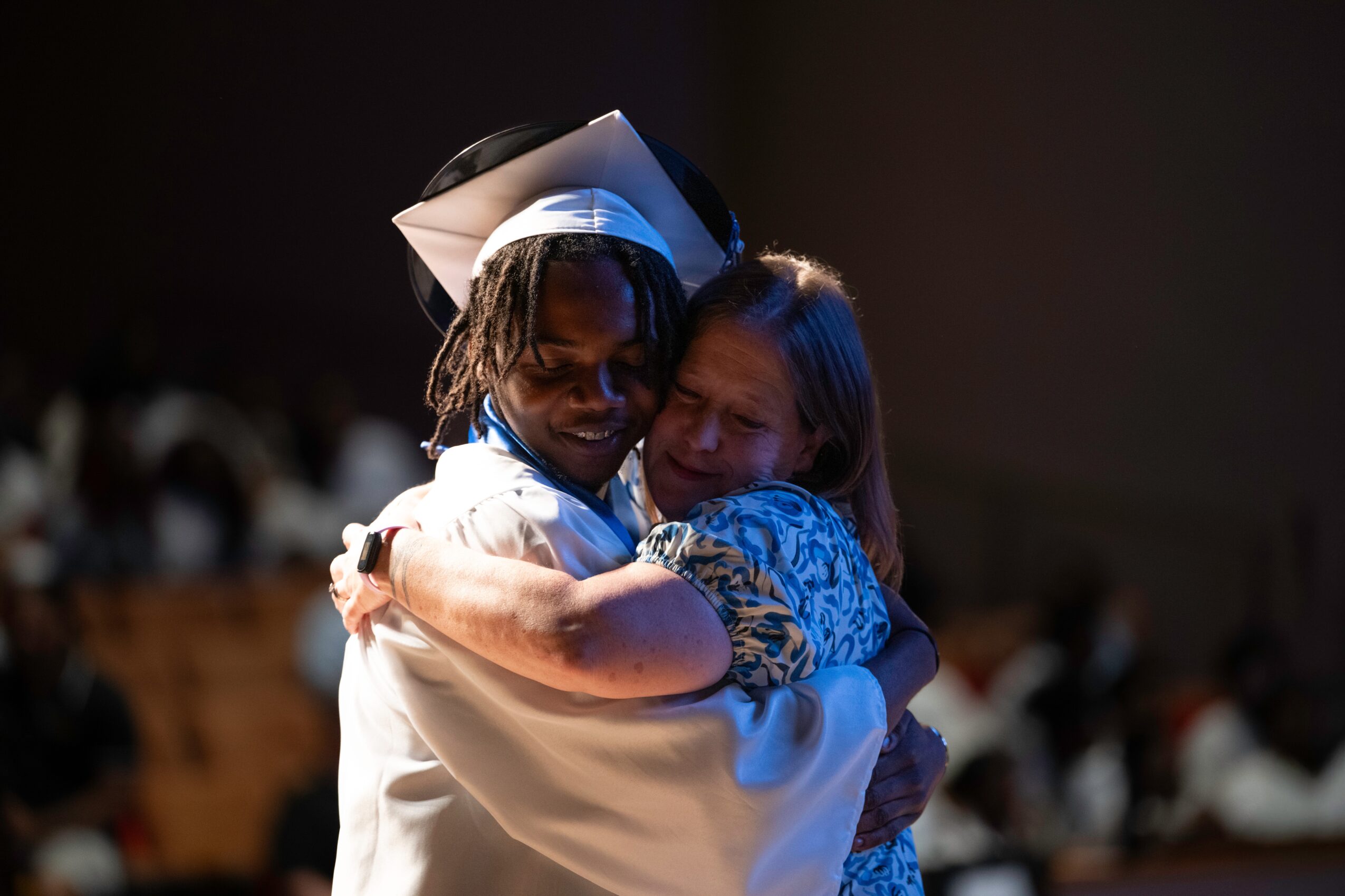 Two people hugging, one wearing a graduation cap, in a dimly lit setting with blurred background.
