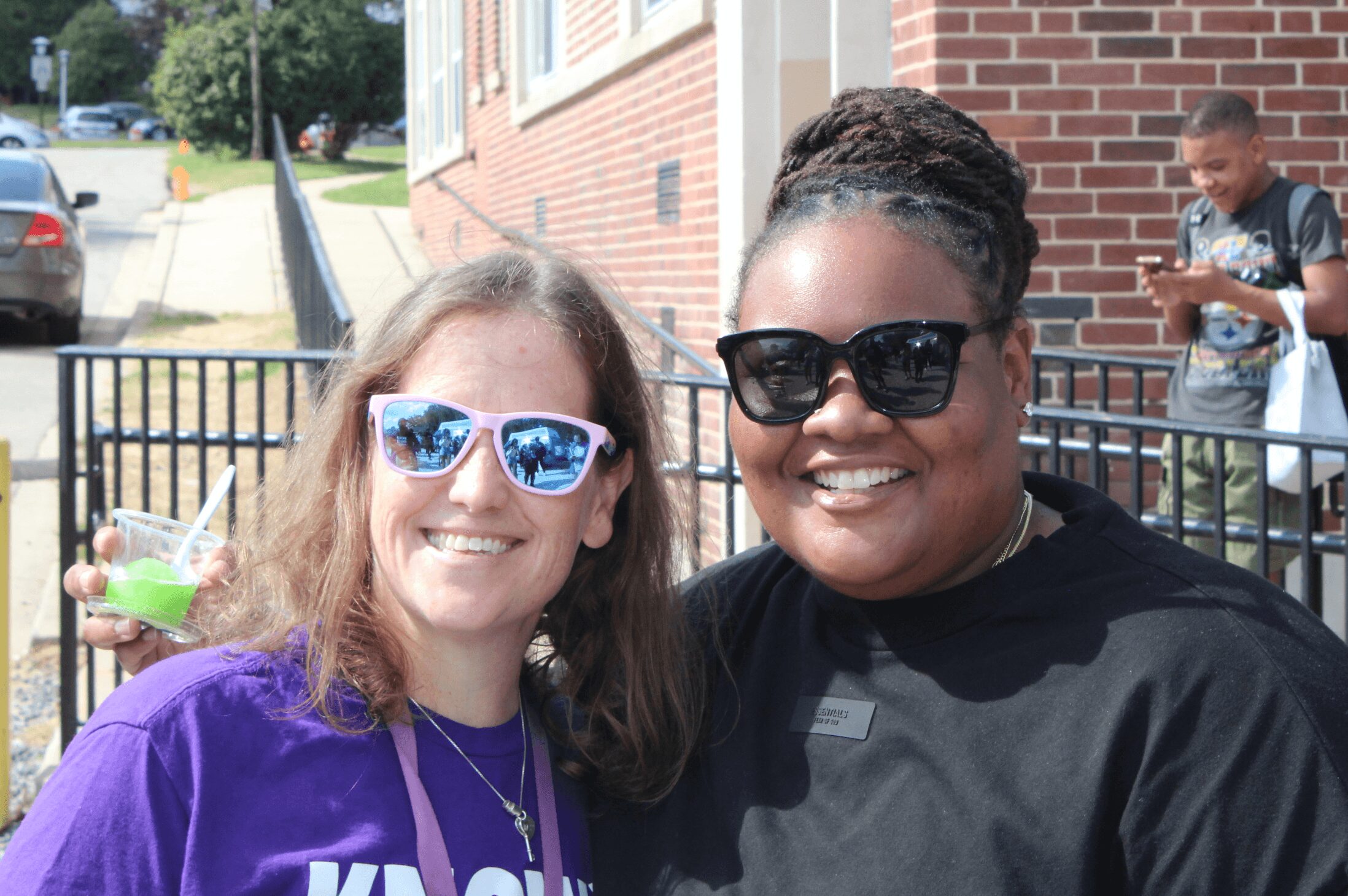 Two women smiling outdoors, one with sunglasses and curly hair, the other with dark sunglasses and braided hair, in front of a brick building.