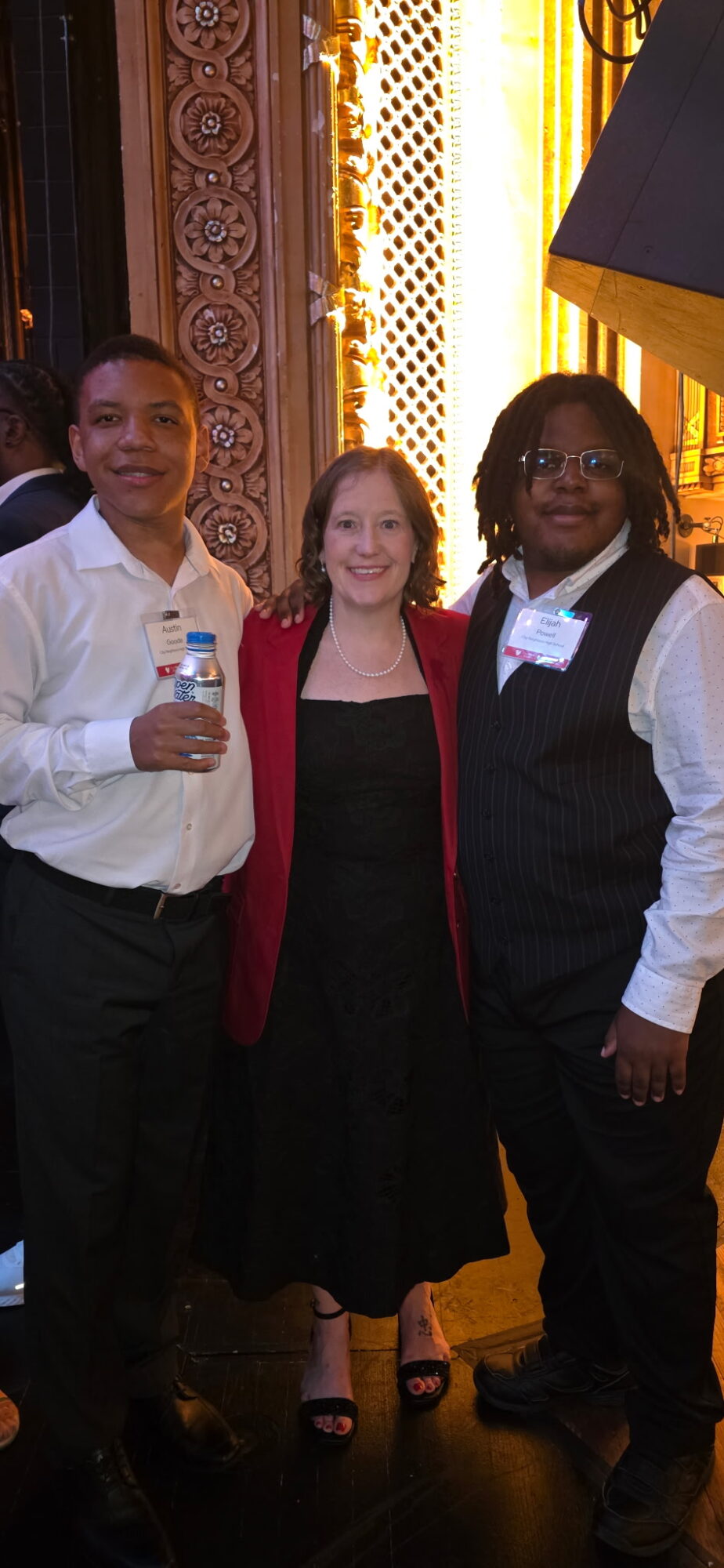 Three people standing together indoors, smiling, with ornate decor and warm lighting in the background.