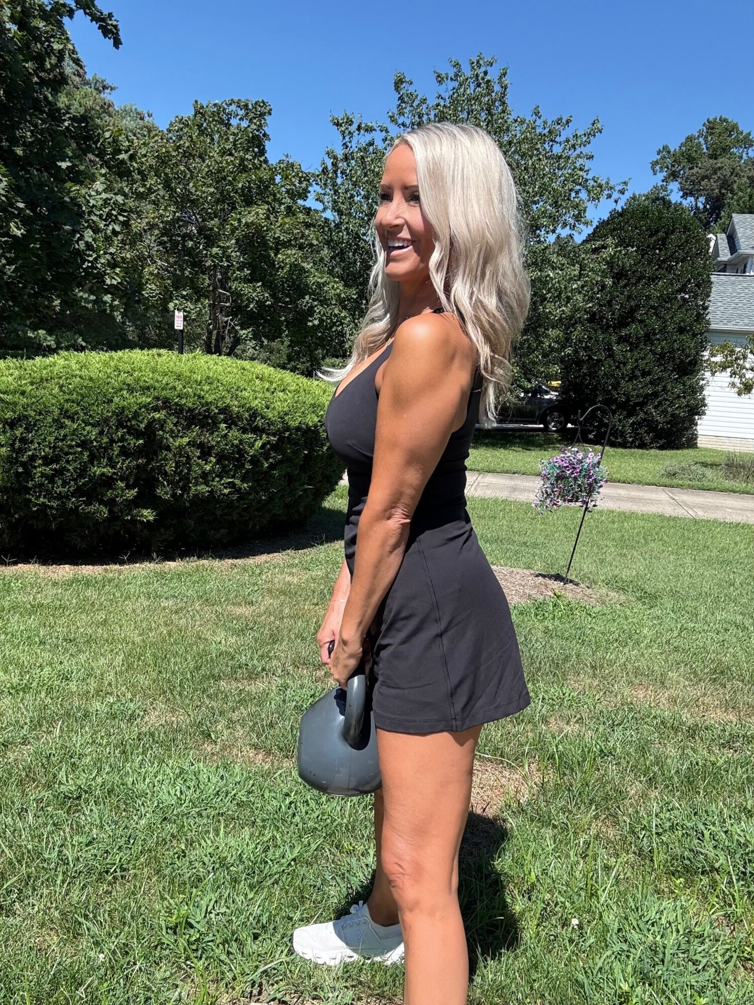 Woman with long blonde hair in a black sleeveless dress holding a gray hat outdoors on grass, smiling, trees, and blue sky in background.