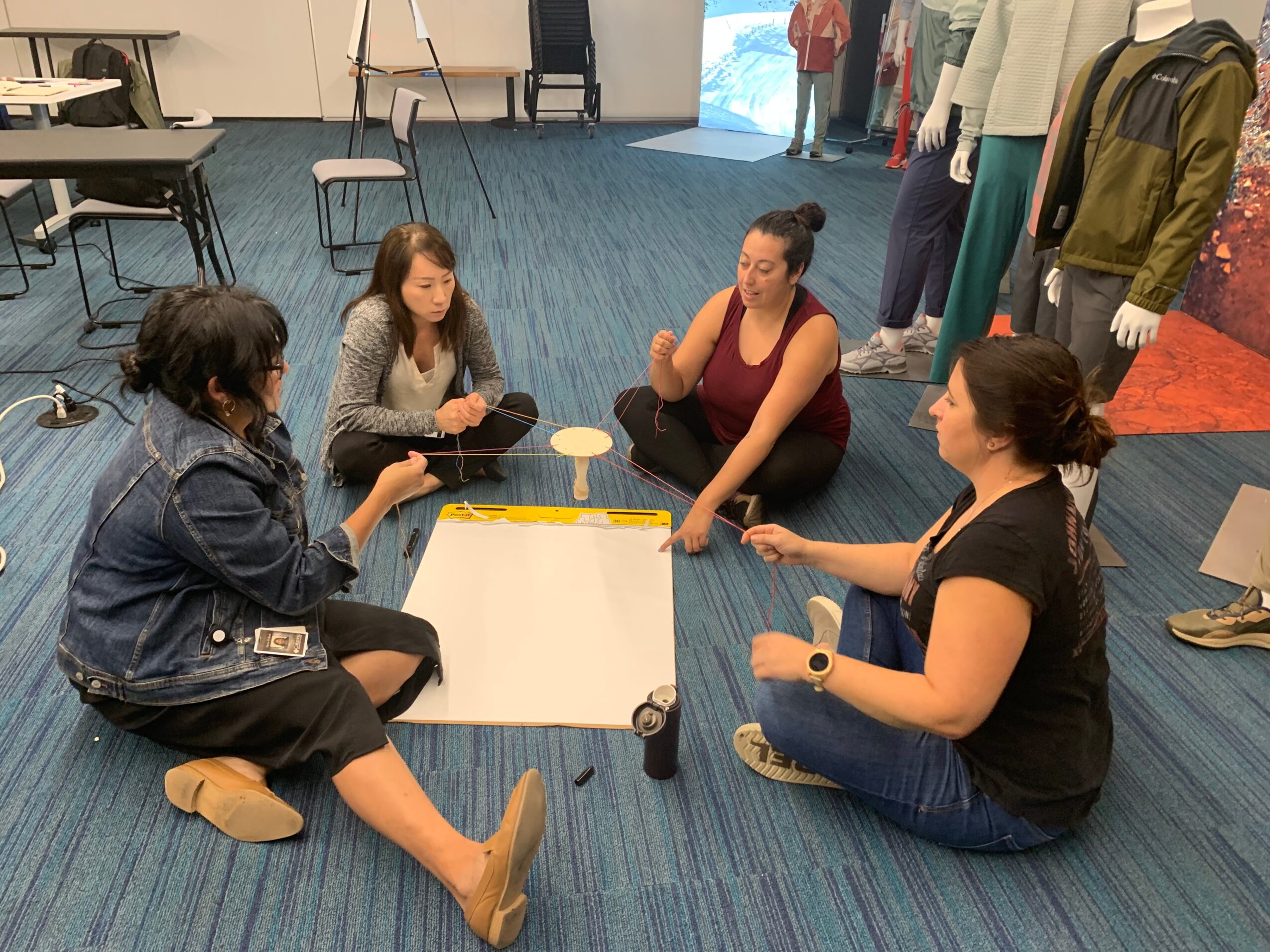 Four women sit on a blue carpeted floor playing a game with a whiteboard and markers, in a room with chairs and mannequins.