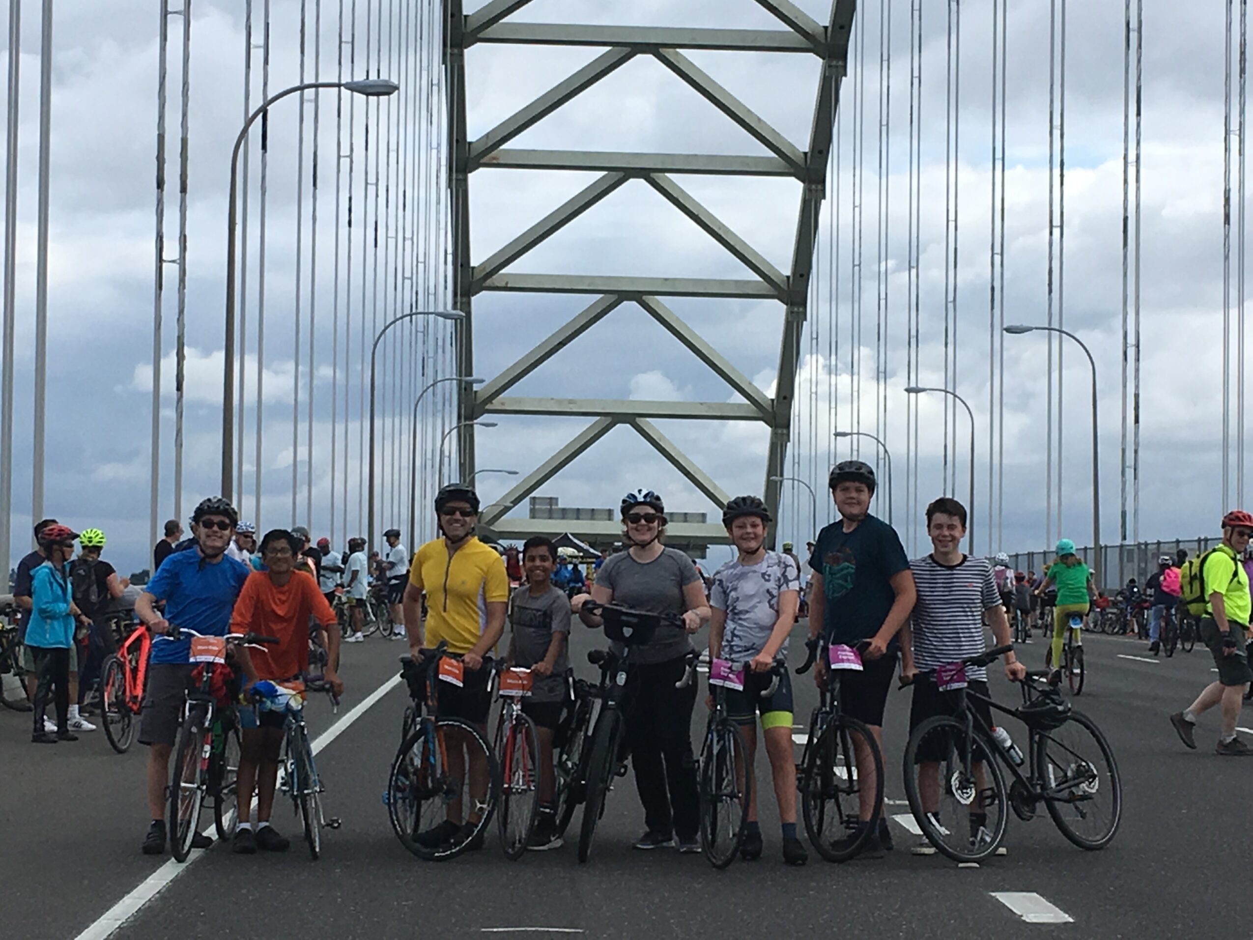 Group of people with bicycles standing on a bridge, with a large metal structure overhead and cloudy sky.