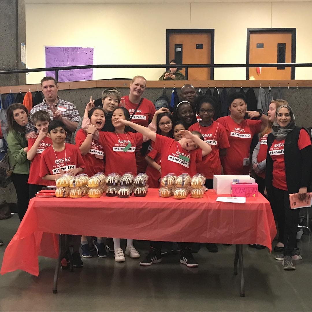 Group of children and adults standing behind a table with cupcakes, in a room with beige walls and two doors, smiling and posing.