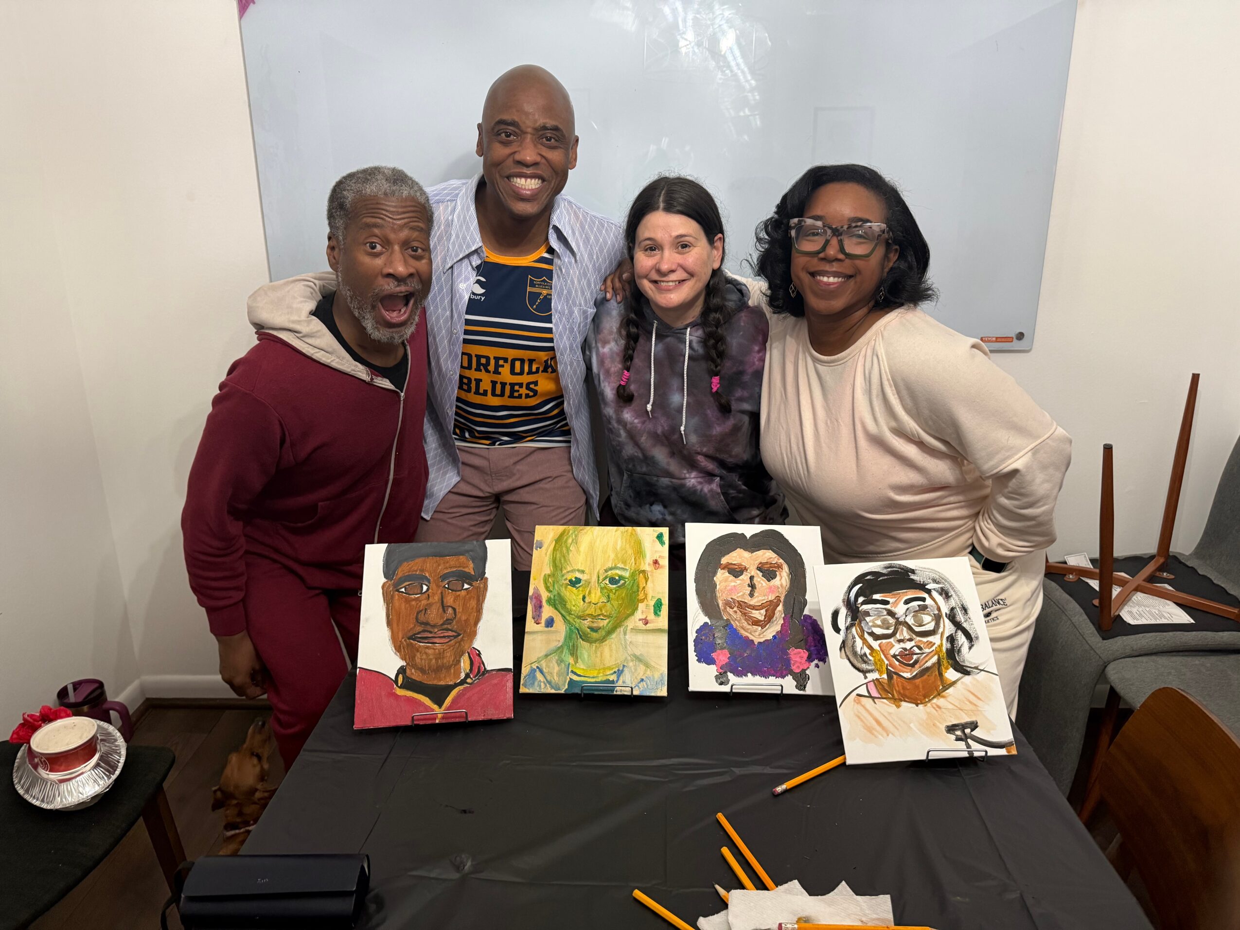 Four people stand behind a table with four colorful portrait paintings, smiling in a room with a whiteboard.