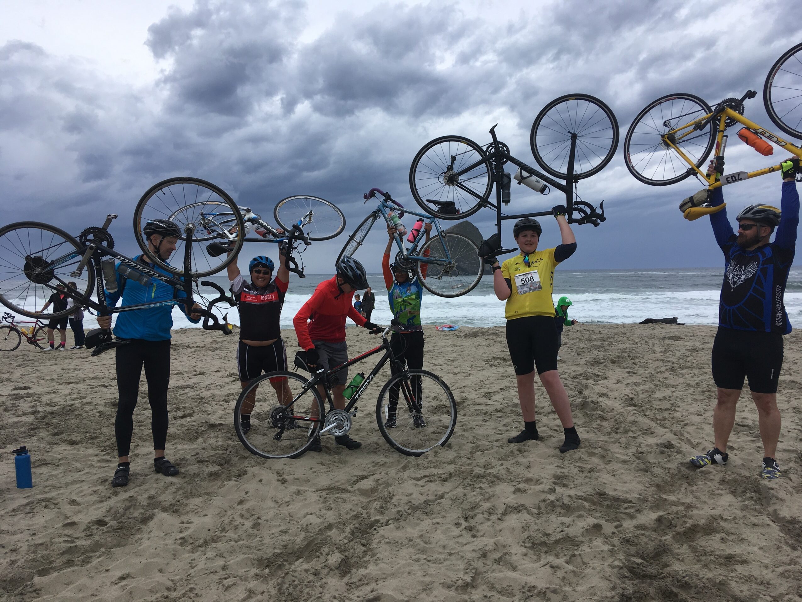 Group of people holding bicycles above their heads on a sandy beach with cloudy sky and ocean in background.