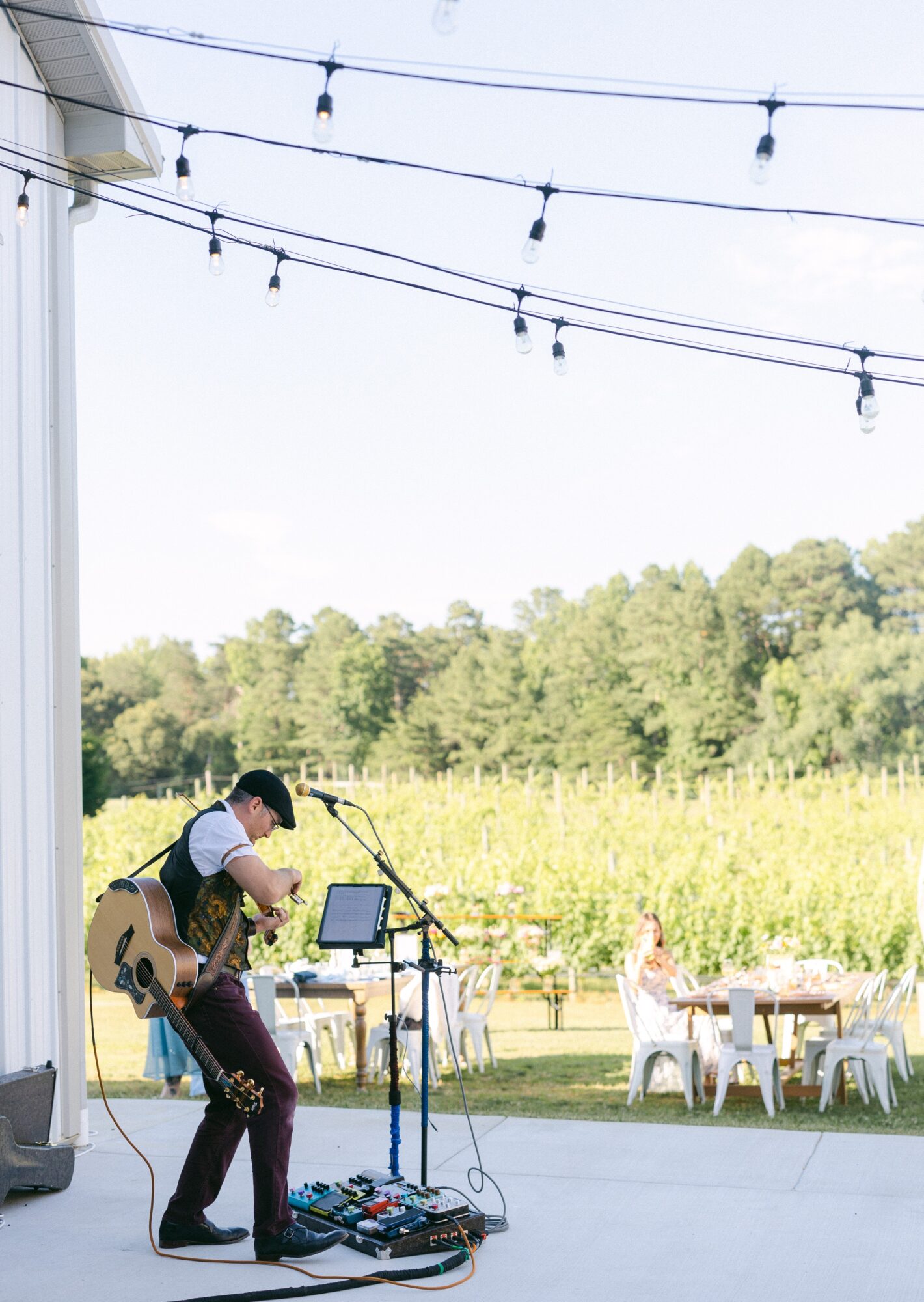Musician playing guitar outdoors with trees and tables in background, string lights overhead, equipment on stage, bright sky.