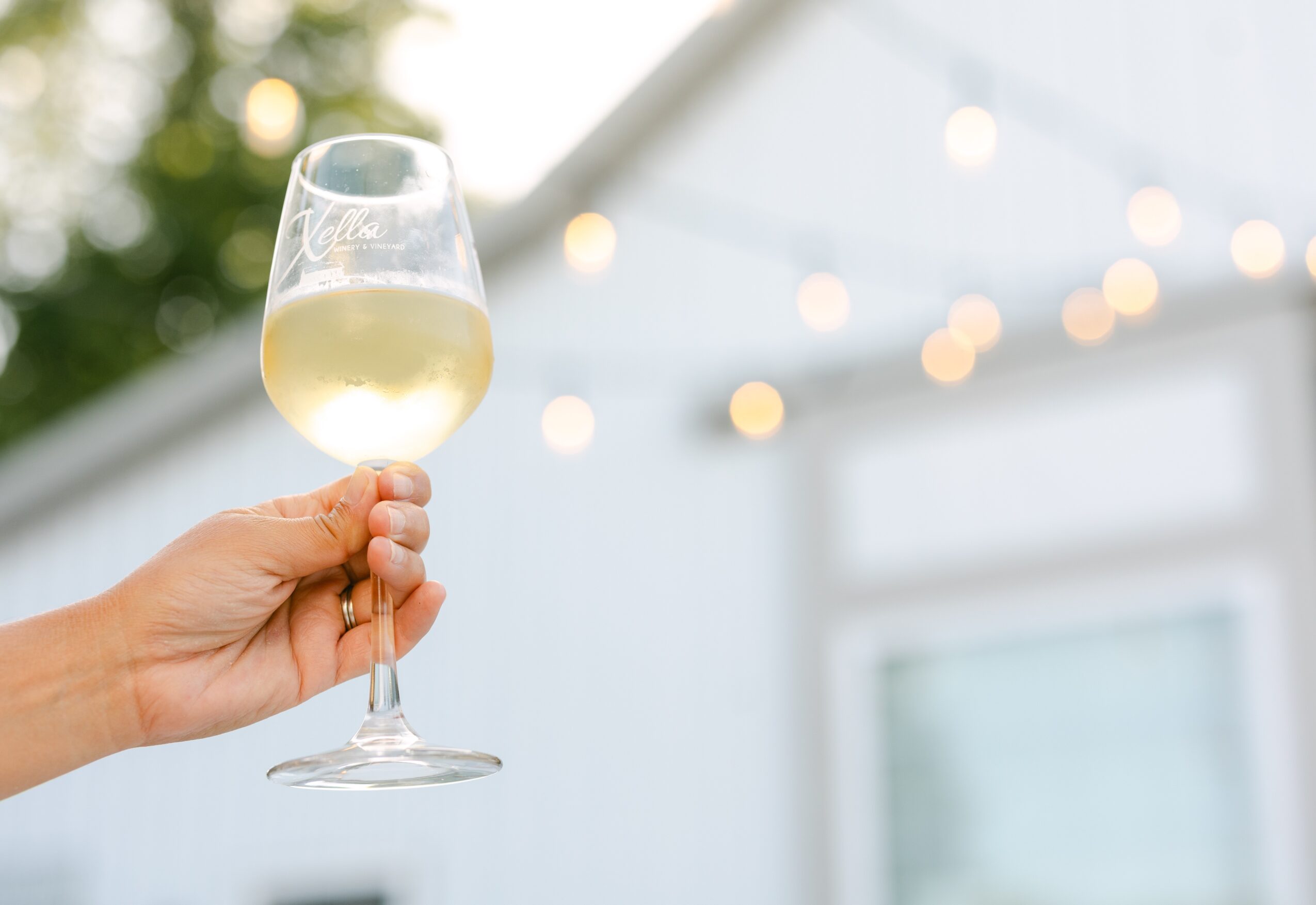 Hand holding a glass of white wine against a background with string lights and a building window.