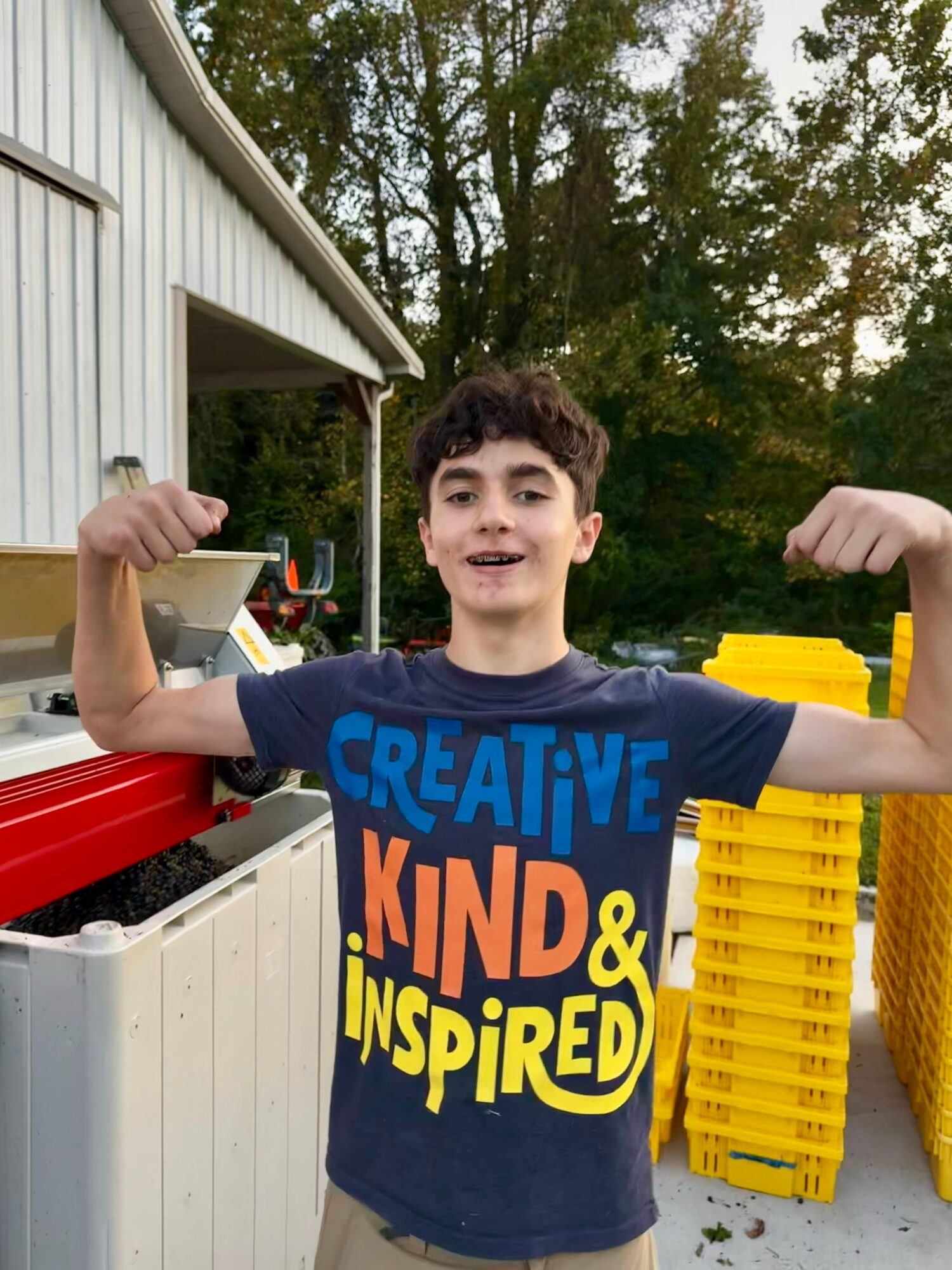 Young boy outdoors flexing arms, smiling, wearing a colorful shirt, with trees and yellow crates in background.