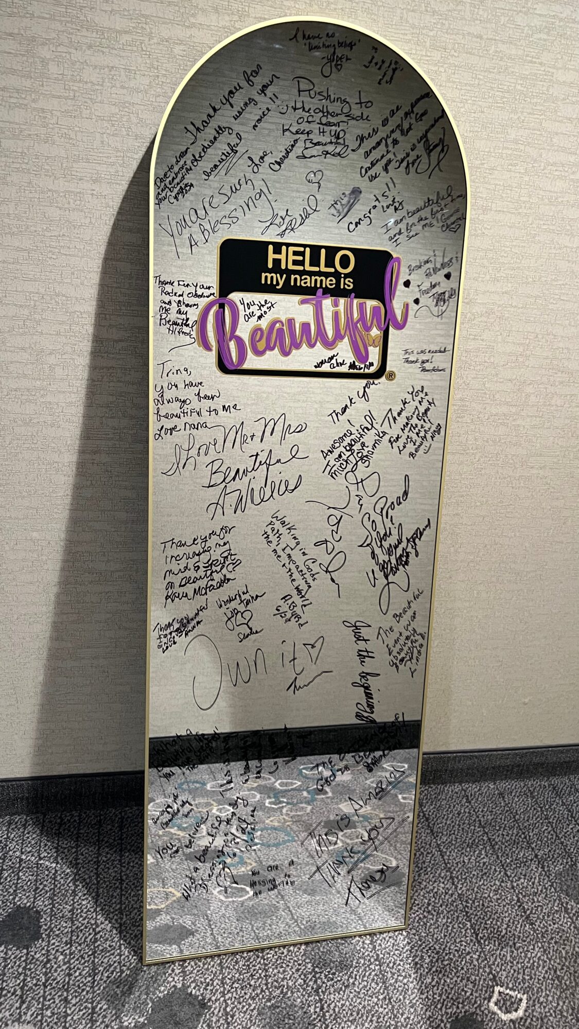 Mirror with handwritten messages and a purple and yellow sign that says 'HELLO my name is Beautiful'.