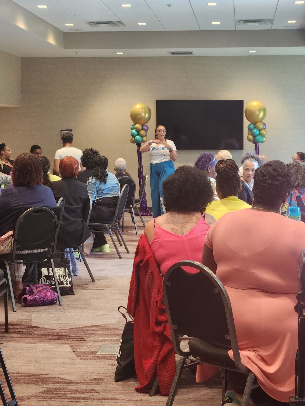 Woman speaking on stage in front of seated audience in a conference room with balloons, TV screen, and ceiling lights.