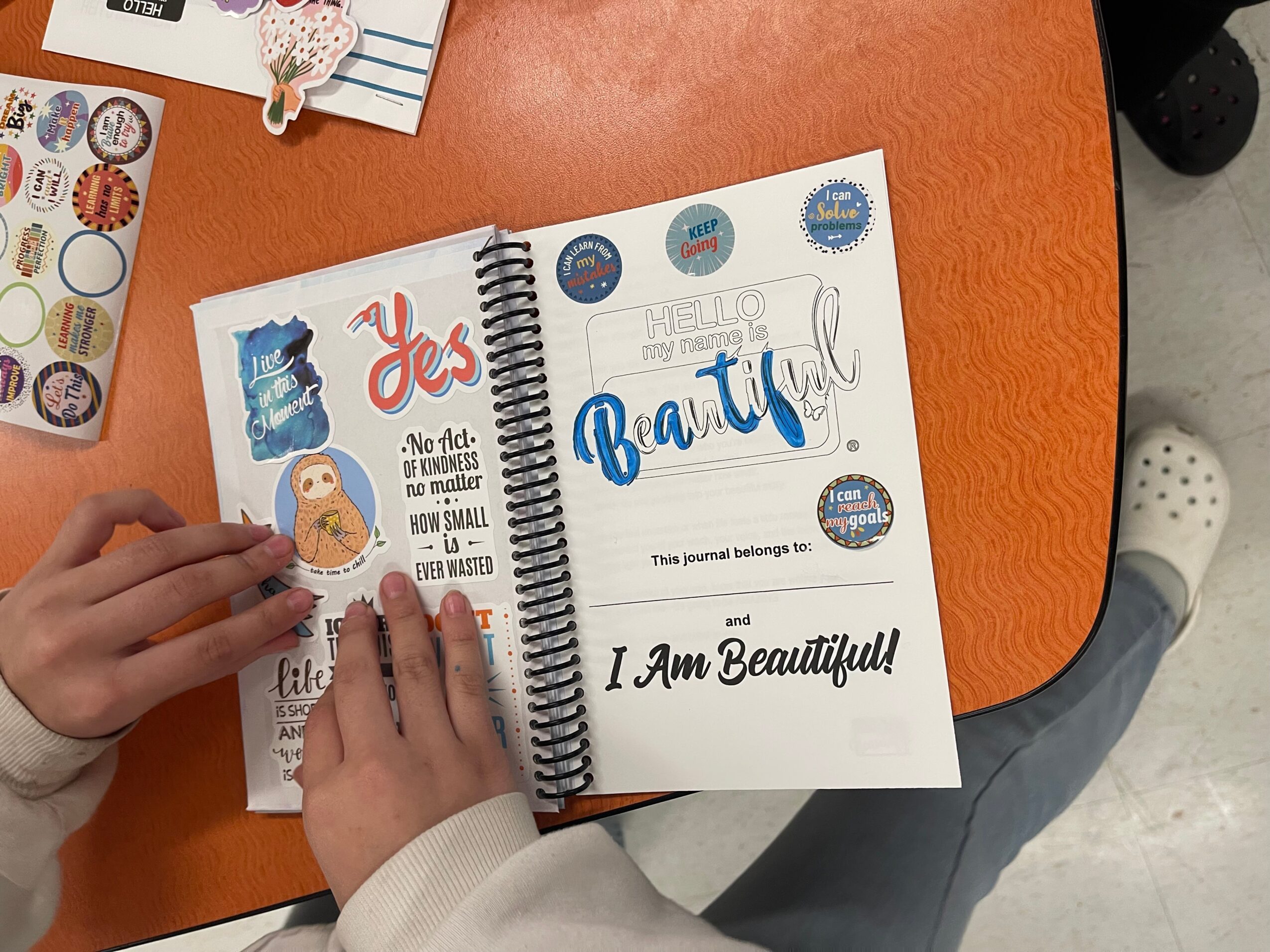 Open journal with decorative stickers and handwritten text, on orange table, person wearing white sweater and gray pants.