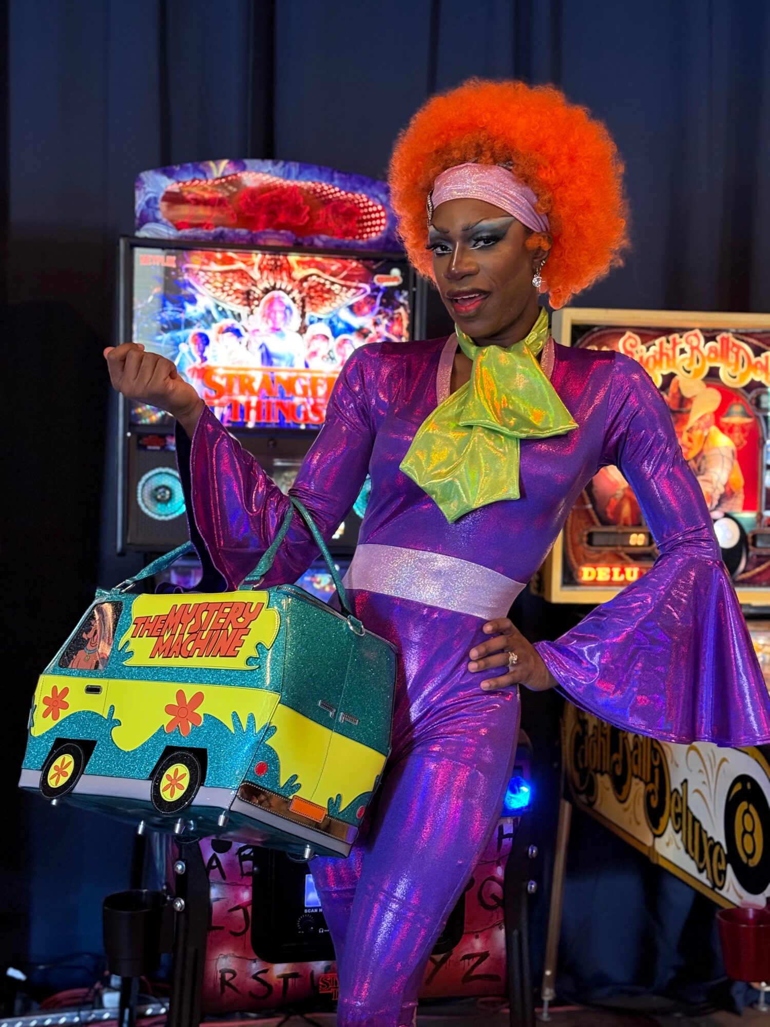 Person dressed in colorful costume with orange wig, standing in front of arcade games, holding a decorated box, smiling.