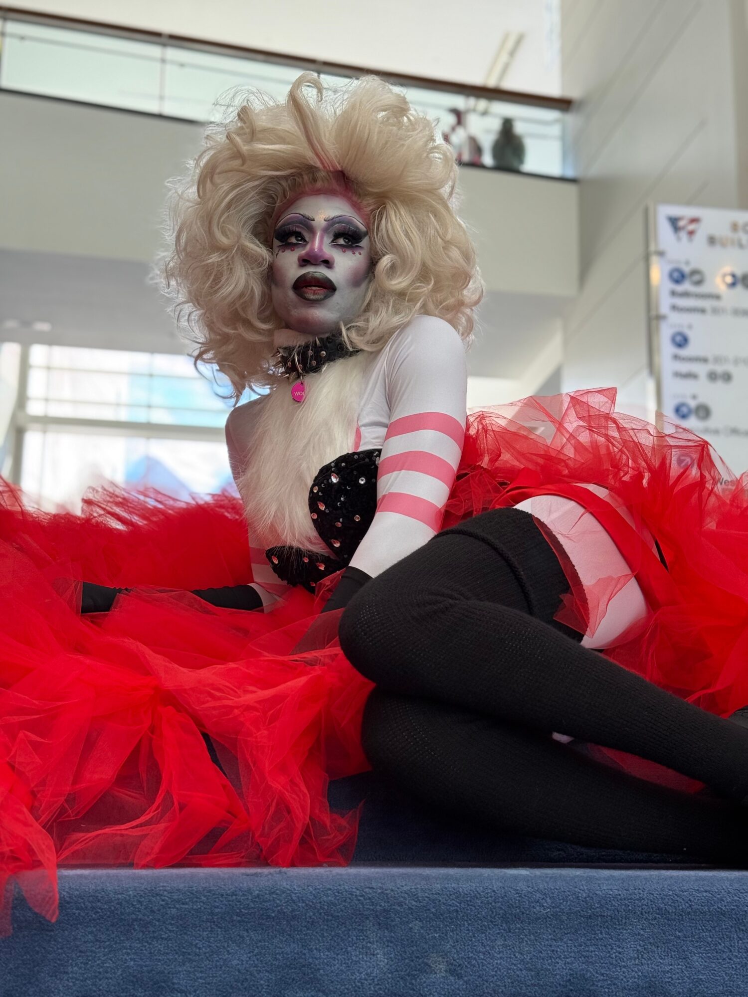 Person with elaborate makeup and hair, wearing a red and black outfit with striped arm warmers, sitting on a surface.
