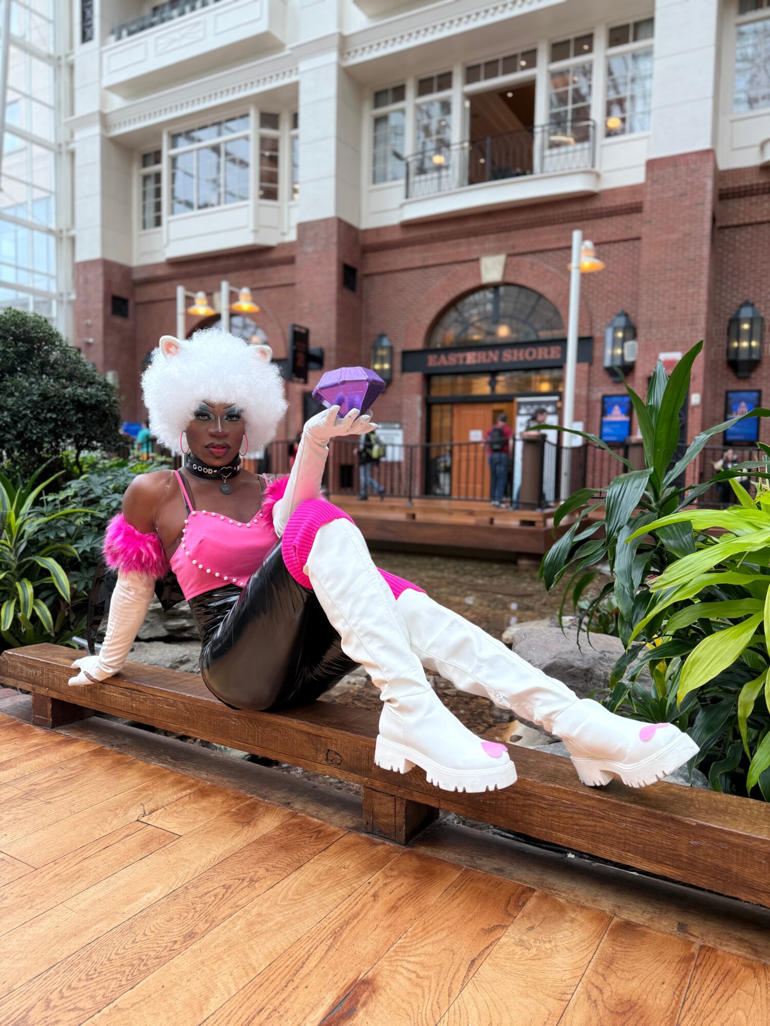 Person with white curly hair, pink top, white thigh-high boots, sitting on a wooden bench outdoors, holding a purple object.