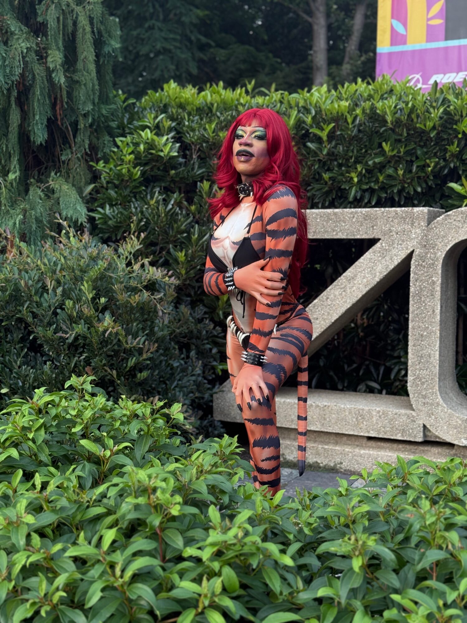 Woman with red hair and striped outfit standing outdoors among greenery and bushes.