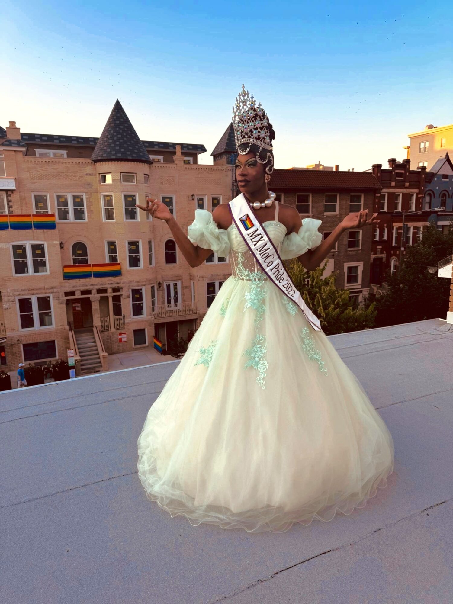Young girl in a light blue dress with a crown and sash, standing on a rooftop with city buildings behind her.