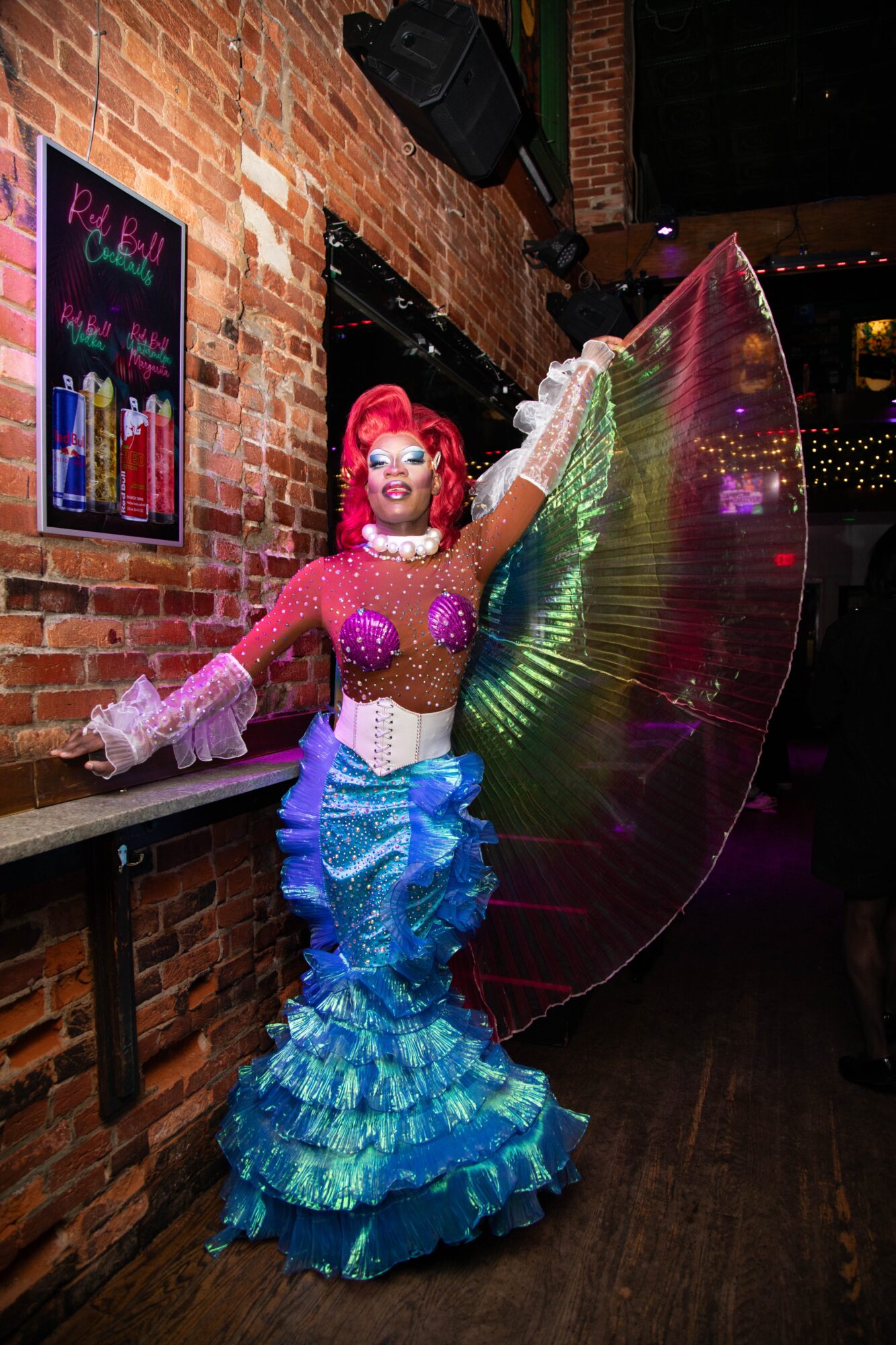 Person in colorful costume with large rainbow fan, standing against brick wall in a dimly lit venue.