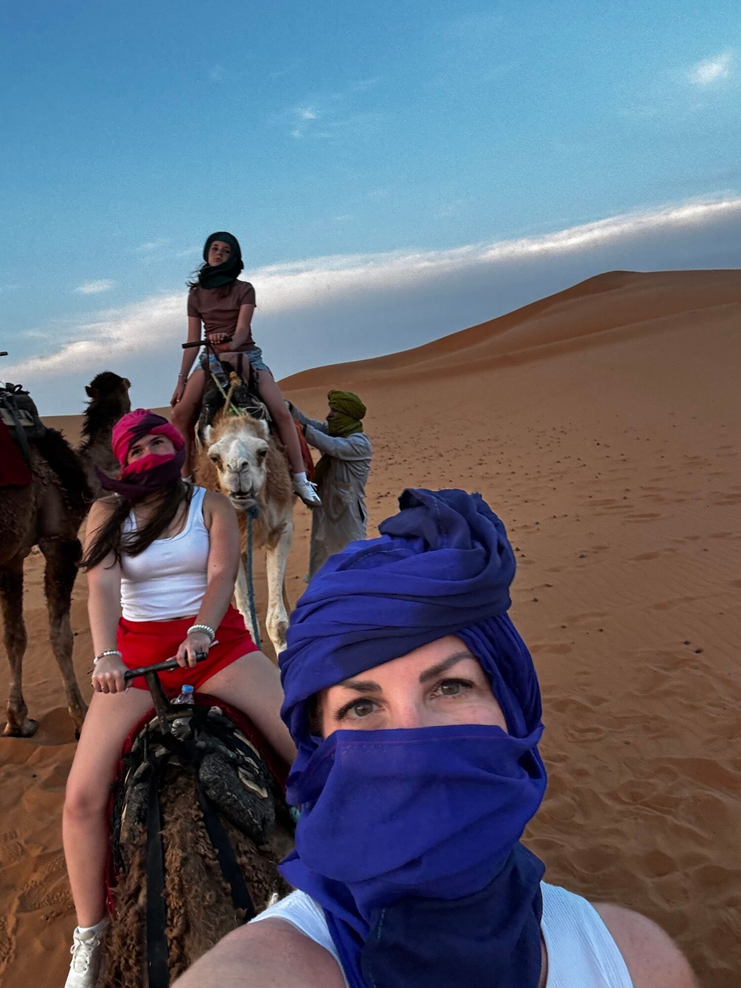 Person with head wrapped in blue cloth taking a selfie on a beach with two women and a man in the background.
