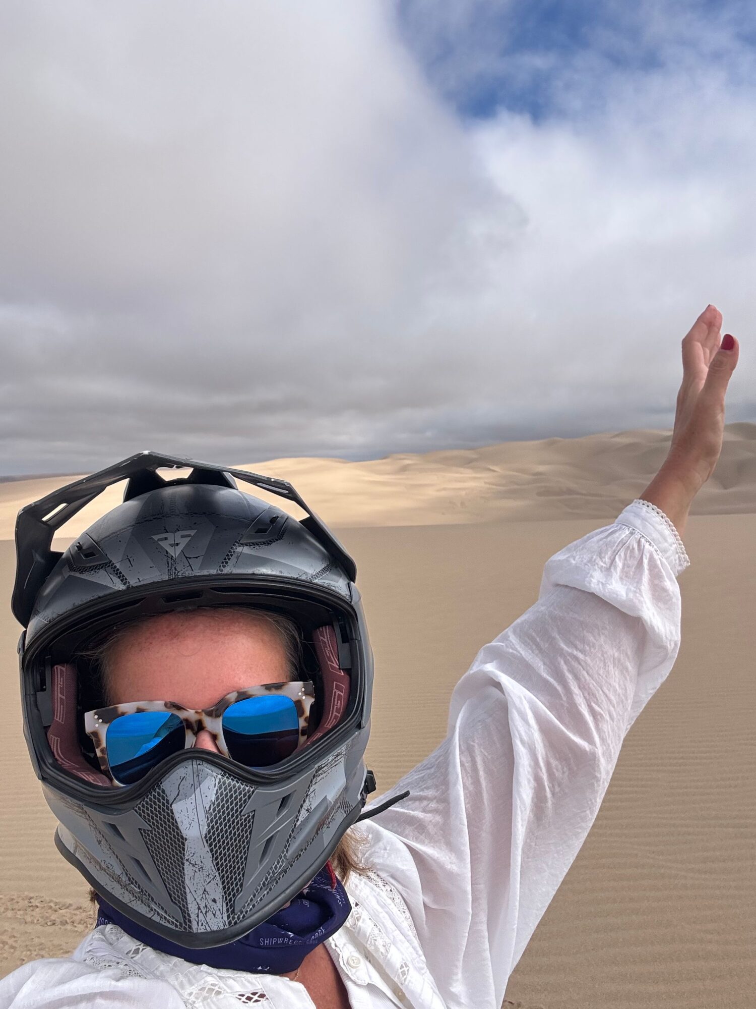 Person wearing a helmet and sunglasses, reaching upward with an arm extended, against a cloudy sky background.
