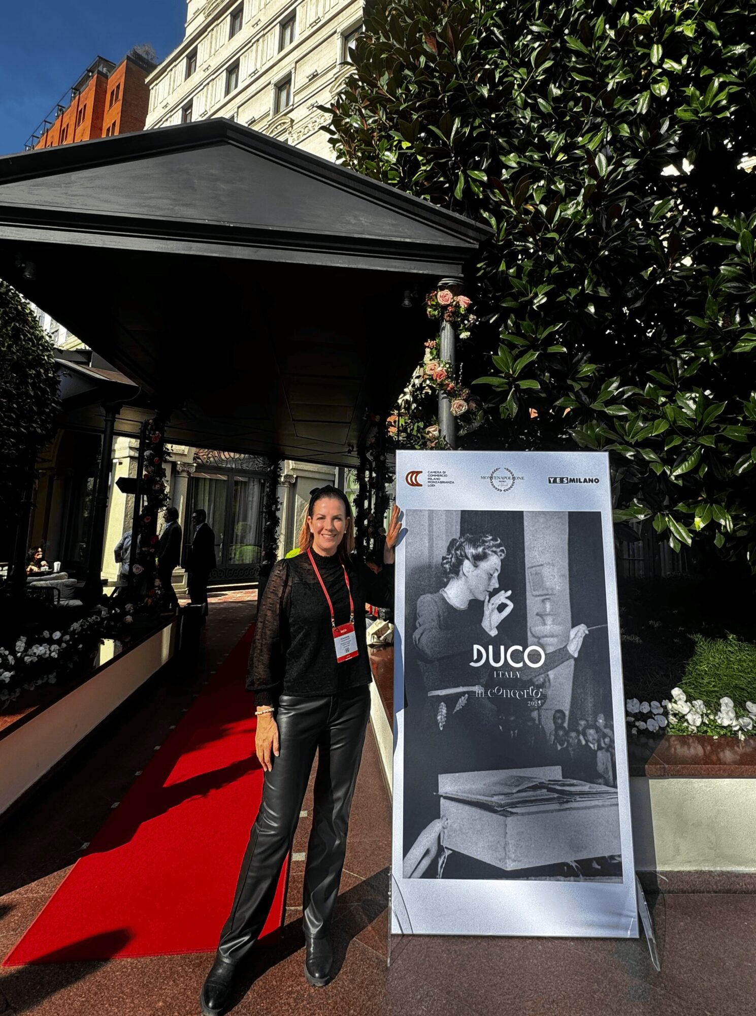 Woman standing on a red carpet outside a building, next to a large poster with a black and white photo, greenery in background.