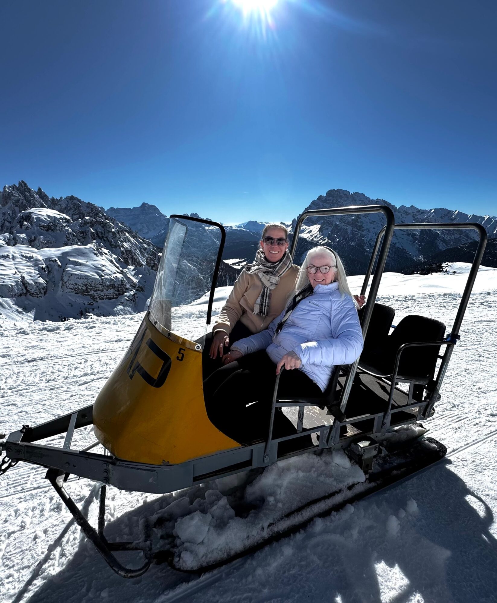 Two people sit on a snow vehicle in a snowy mountain landscape under a bright blue sky.