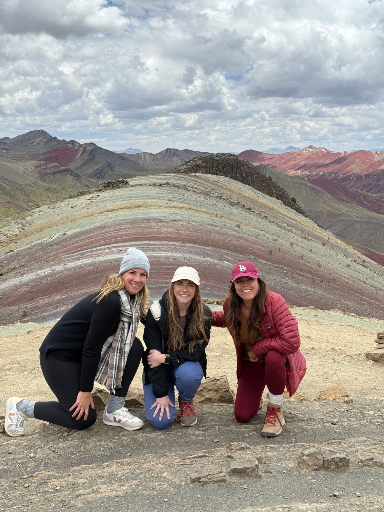 Three women posing in front of colorful layered rock formations, some wearing masks, outdoors on a cloudy day.