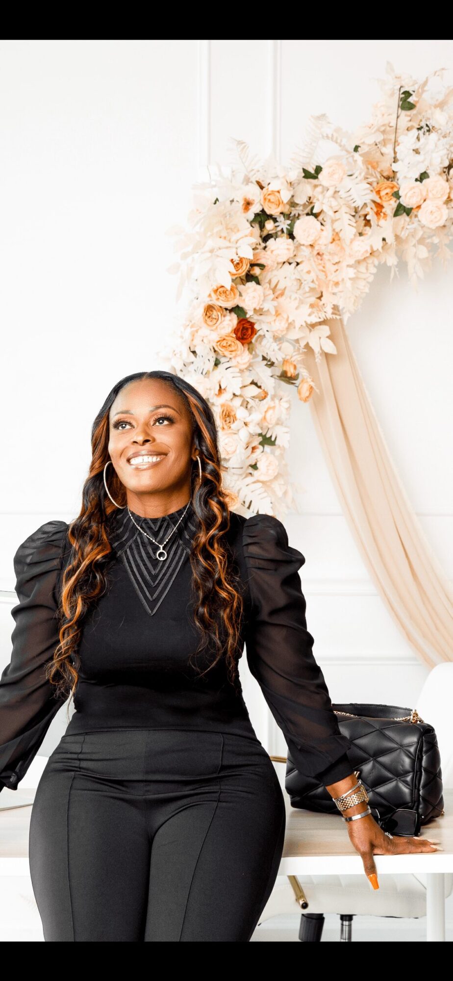 Woman smiling, standing in front of floral arrangement and draped fabric, wearing a black dress with puffed sleeves.