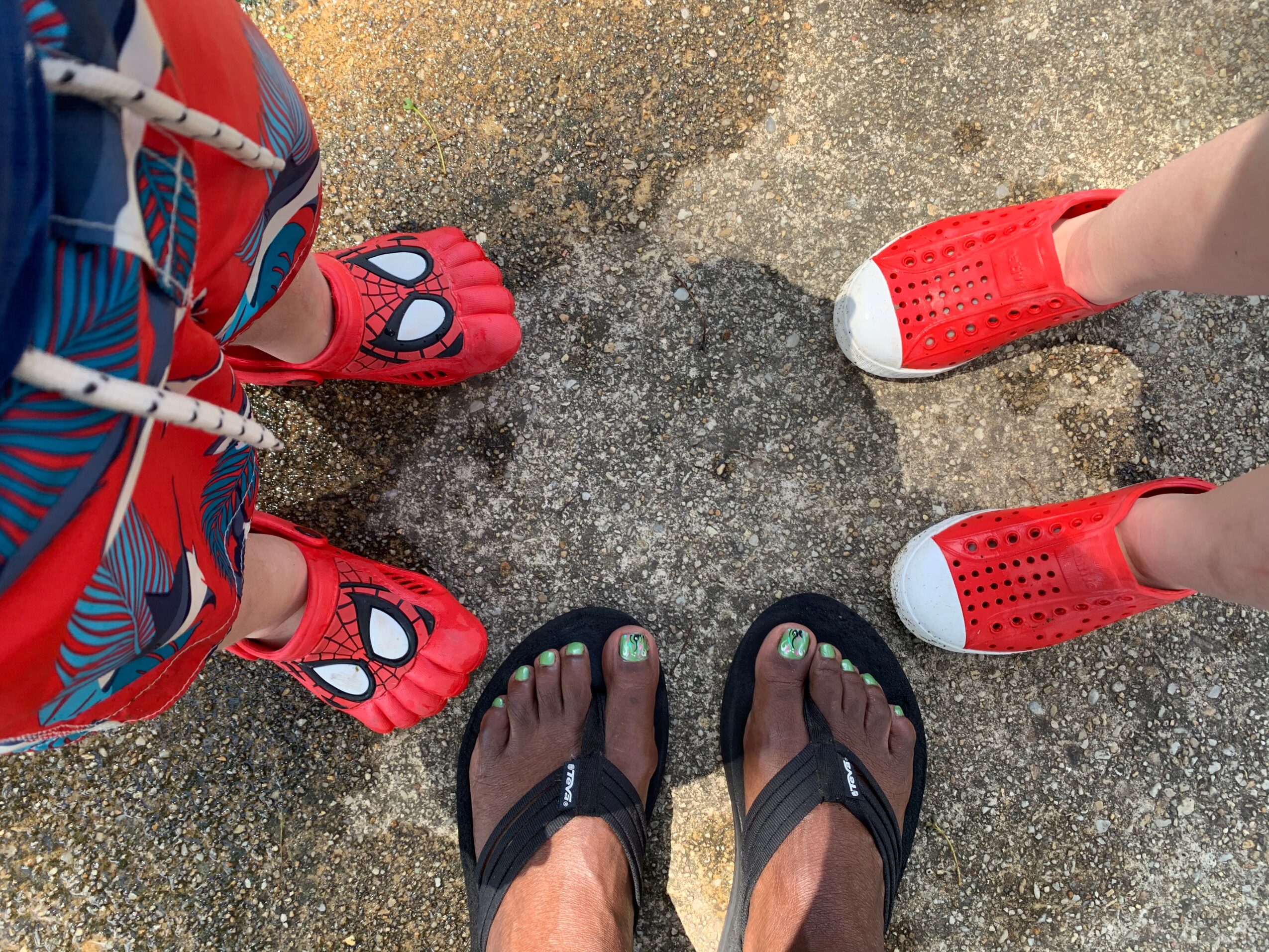 Four people wearing colorful sandals and flip-flops standing on a sandy surface, viewed from above.
