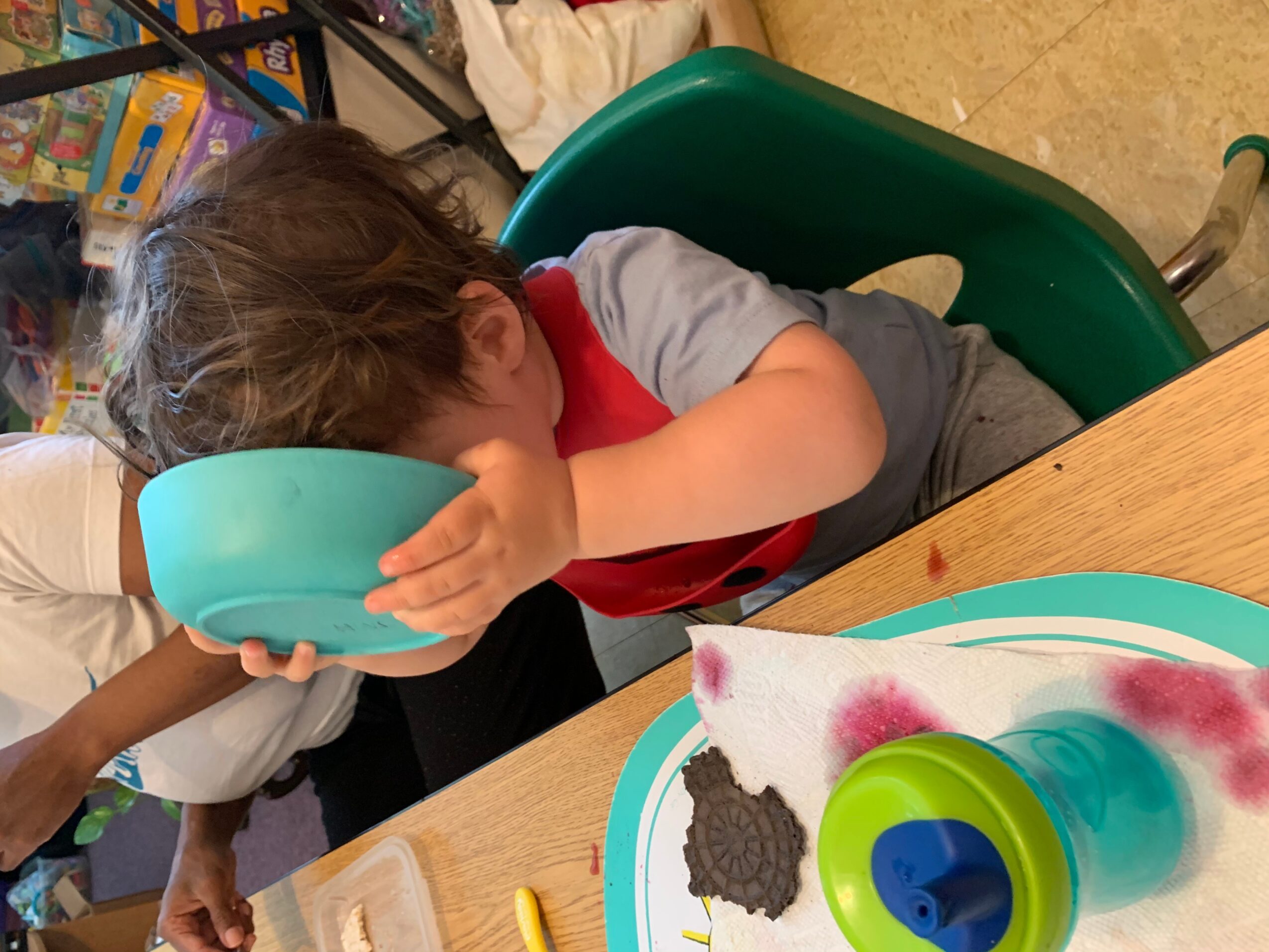 Child with brown hair drinking from a blue bowl, sitting at a table with a colorful tray and a green cup.