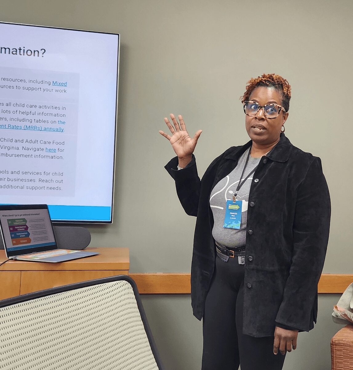 Woman with short curly hair and glasses standing next to a presentation screen, gesturing with her right hand, in a conference room.