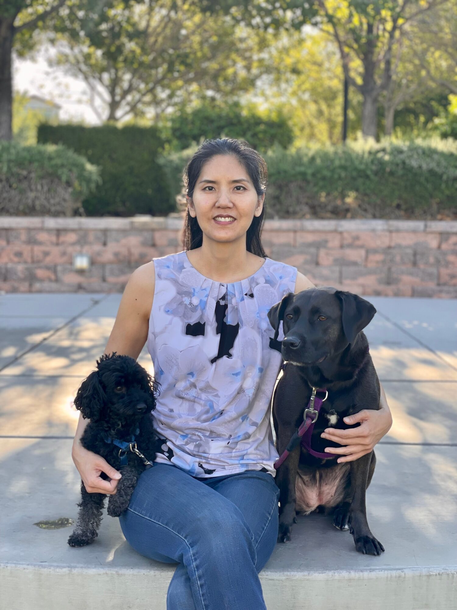 Woman sitting outdoors with two dogs, smiling, trees and bushes in background.