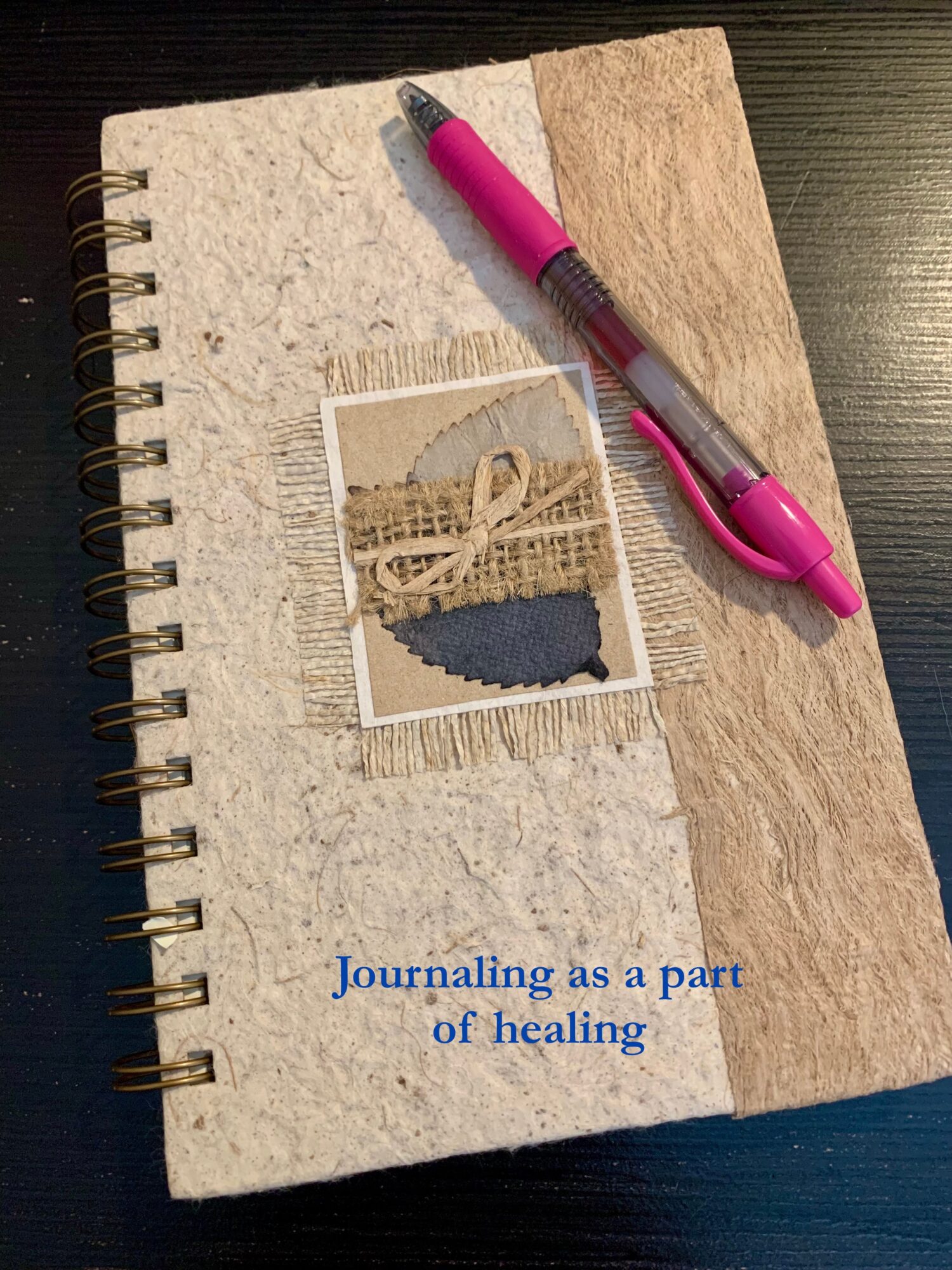 Open spiral notebook with a pink pen on top, a photo, and text reading 'Journaling as a part of healing' on textured paper cover.