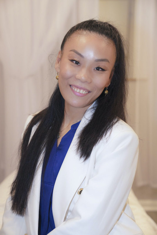 Smiling woman with long dark hair, wearing a white blazer and blue top, in a neutral background.