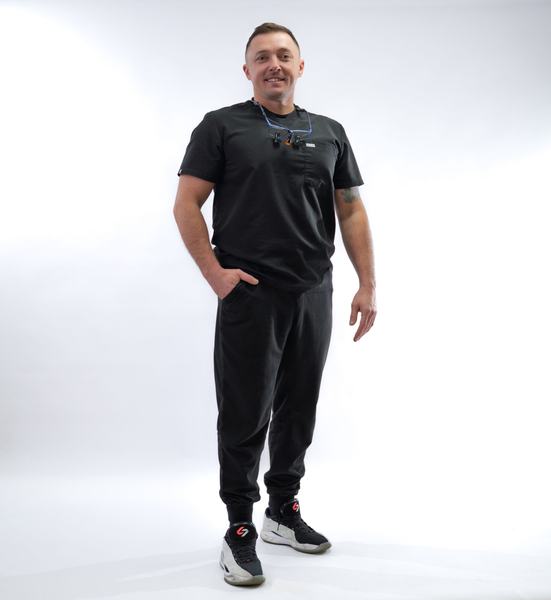 Man in black scrubs standing with one hand in pocket, smiling, against a plain white background.