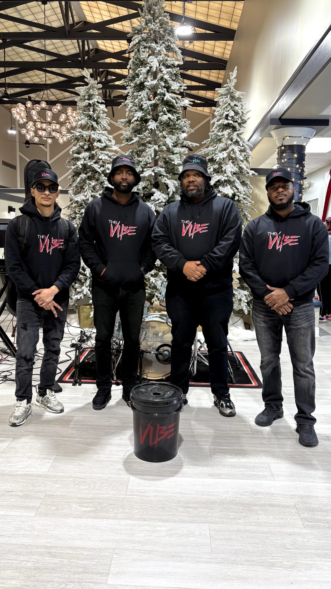 Four men stand in front of decorated Christmas trees in a shopping mall, wearing black hoodies with red text, with a black bucket in front.