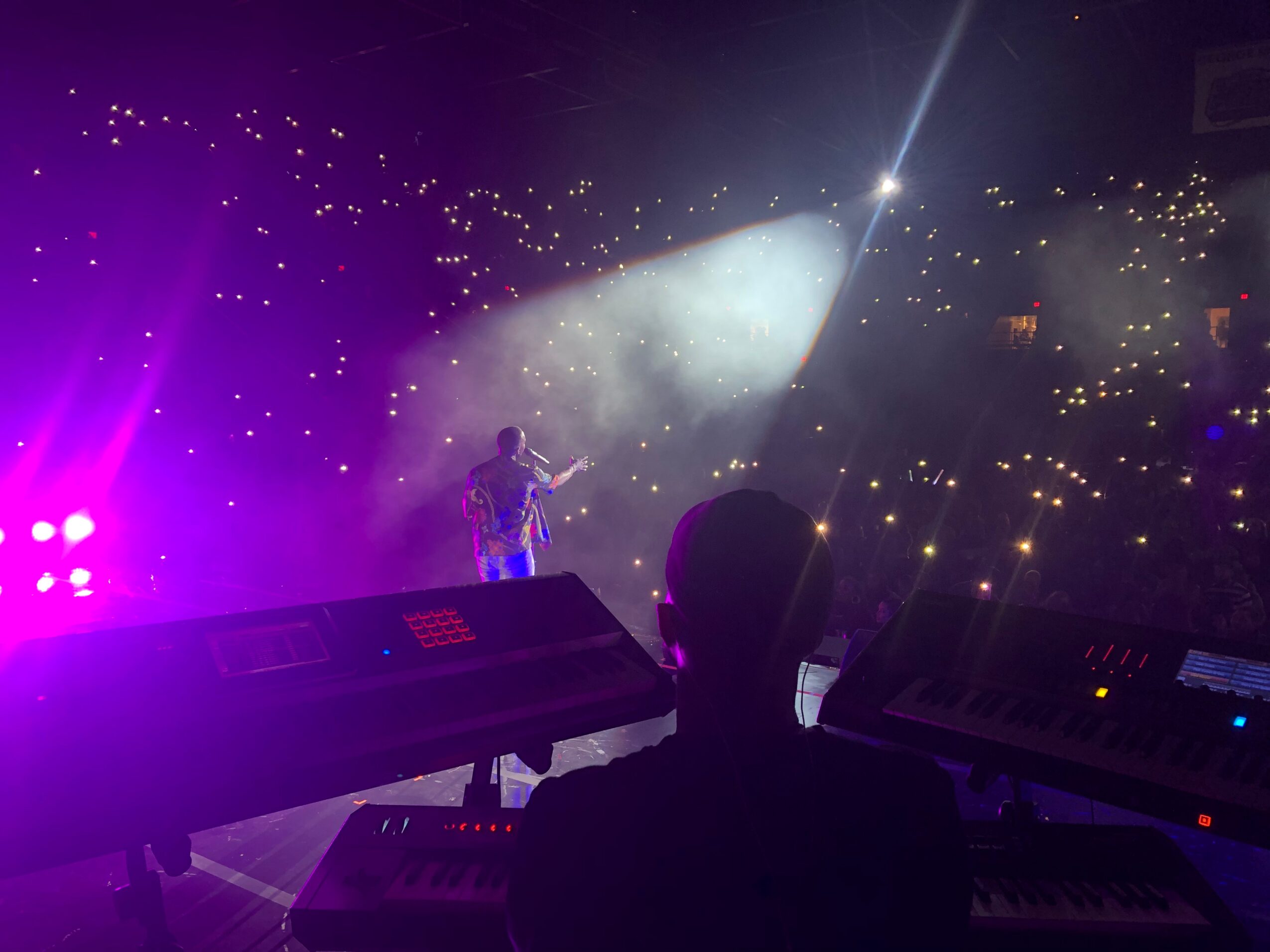 Performer on stage with bright lights and fog, audience holding up lights, silhouette of a person at keyboard, concert atmosphere.