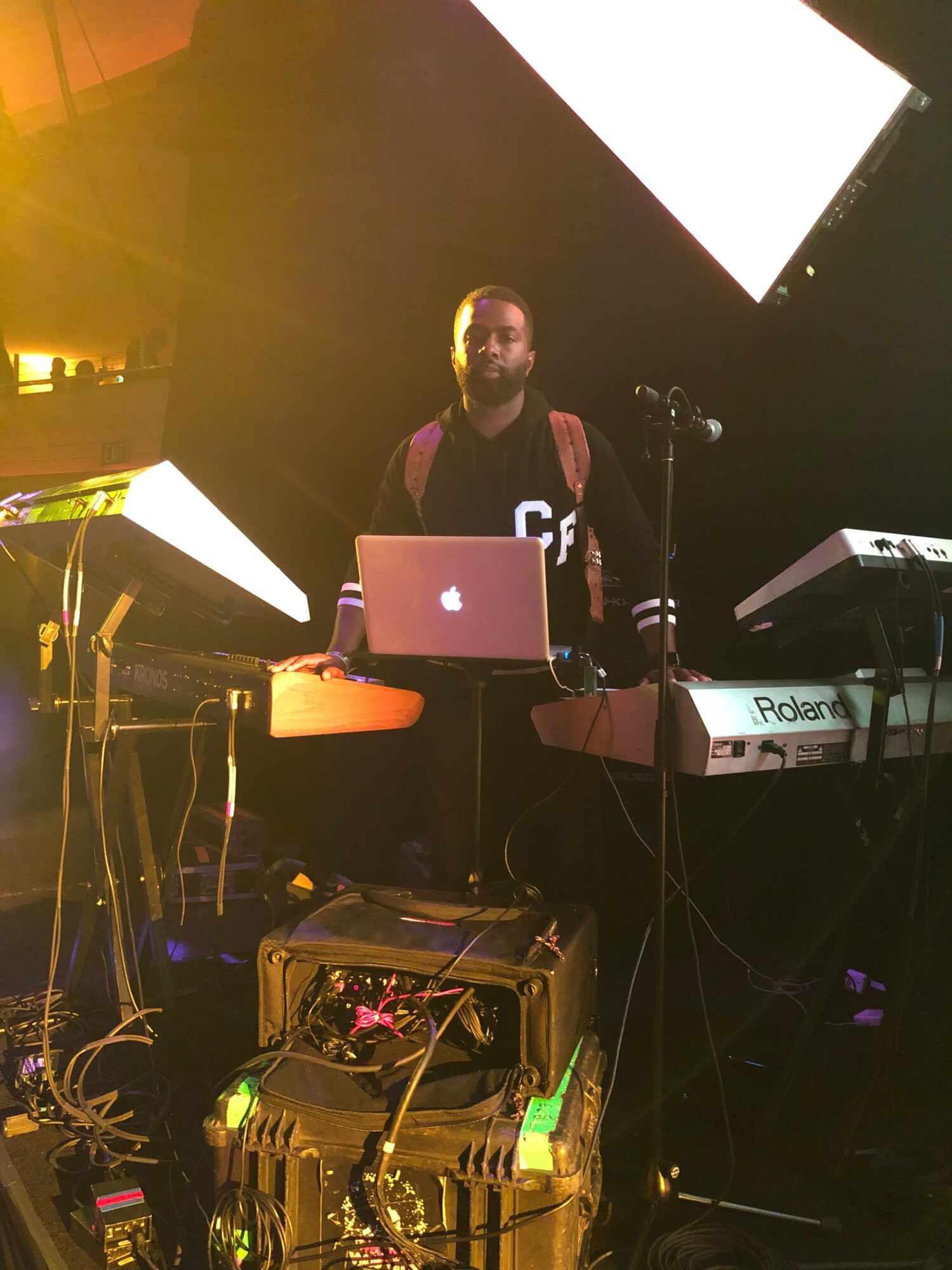 A man with a beard stands behind a laptop and musical equipment on stage, with bright lights and screens above.