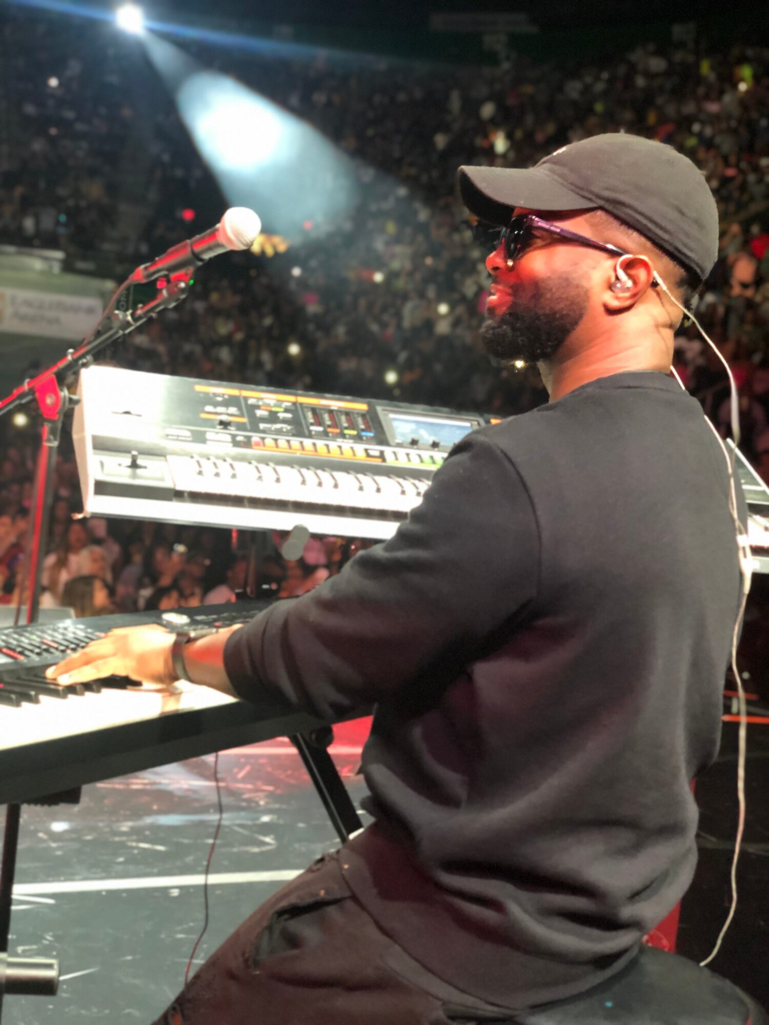 Musician playing keyboard on stage with a crowd in the background, wearing sunglasses and a cap, illuminated by stage lights.
