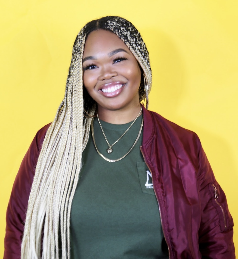 Smiling woman with long blonde braids wearing a green shirt and maroon jacket against yellow background.