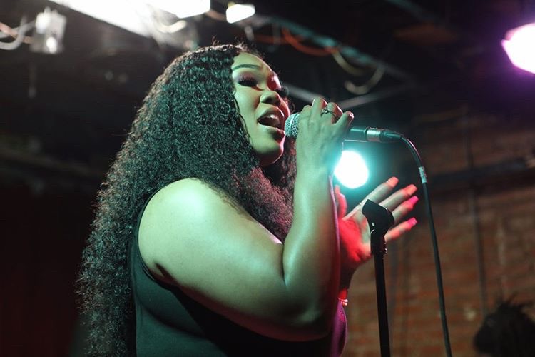 A woman with curly hair singing into a microphone on stage, with stage lights in the background.
