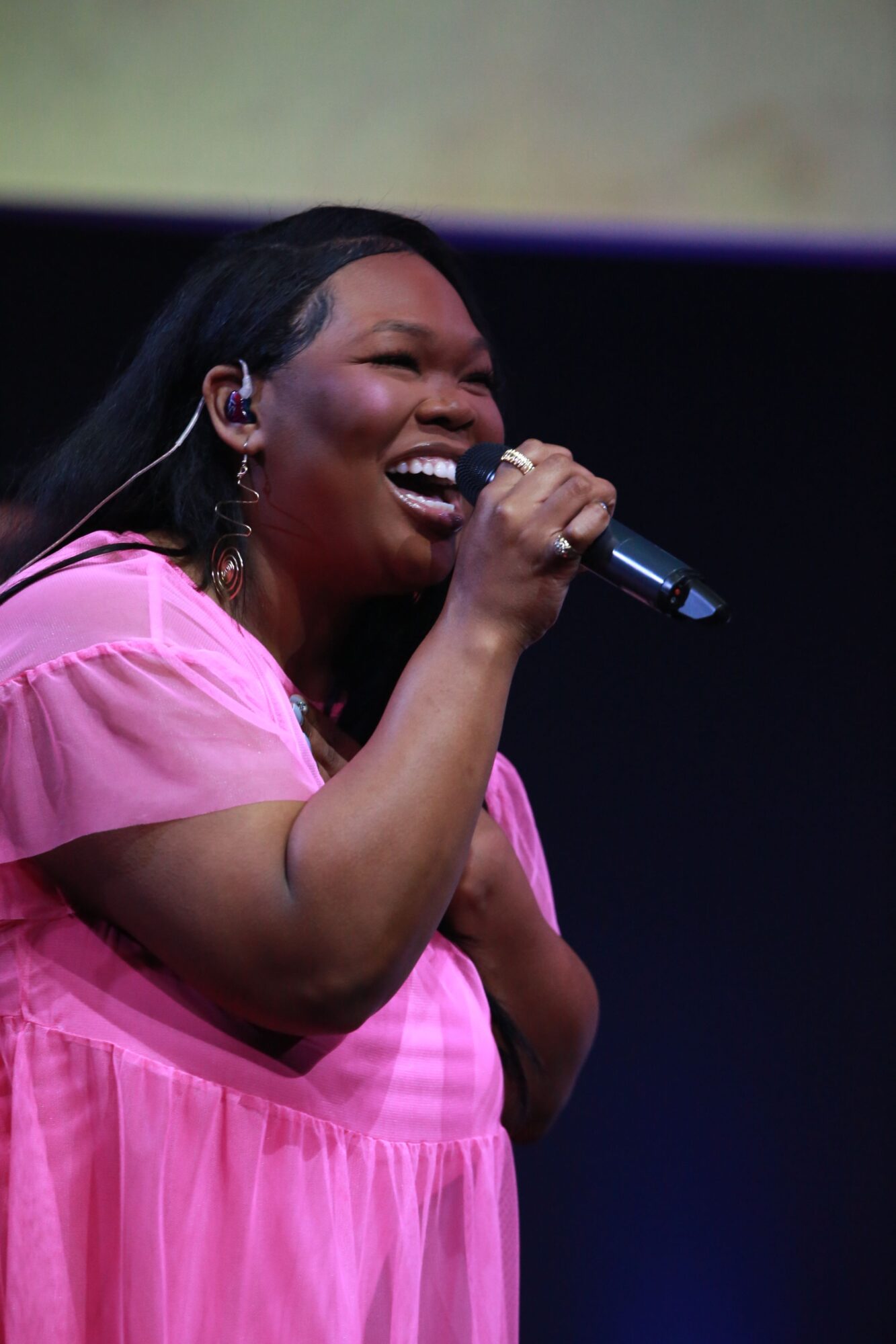 Woman singing into a microphone, smiling, wearing a pink dress, with dark hair and a dark background.