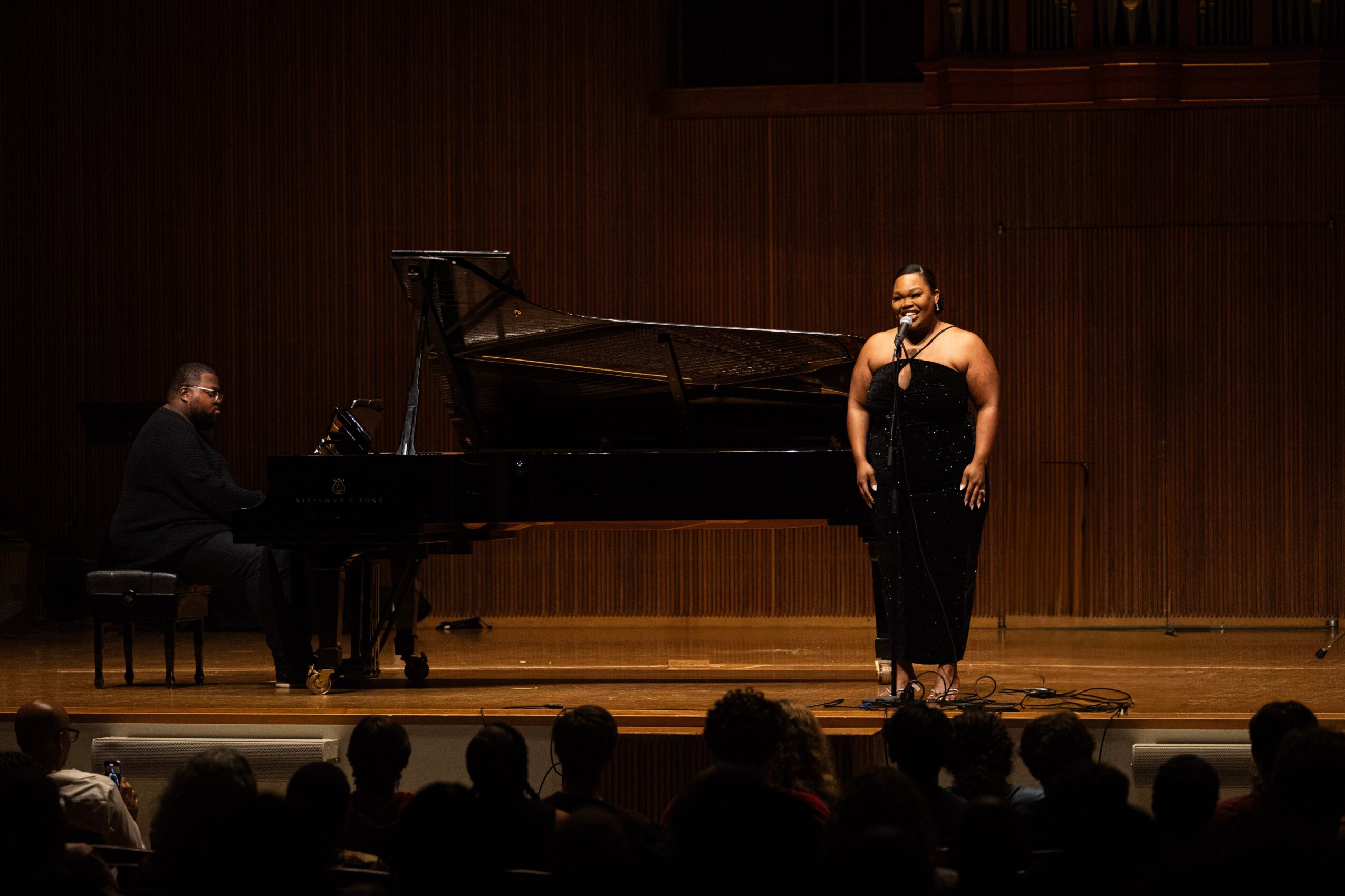 Pianist and singer perform on stage in front of an audience, with a wooden backdrop and stage lighting.