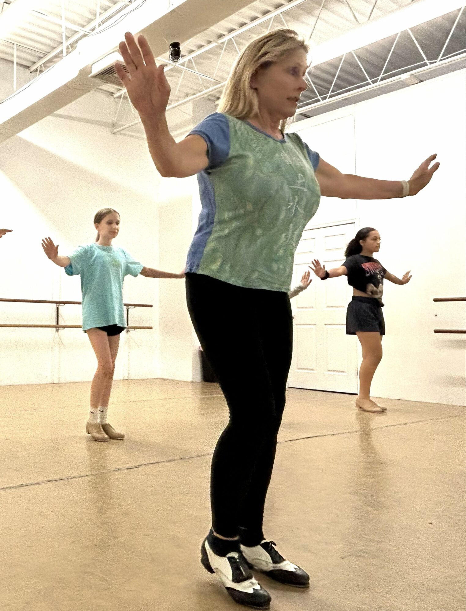 Three women practicing dance or exercise in a studio with wooden floor and high ceiling, arms extended outward.