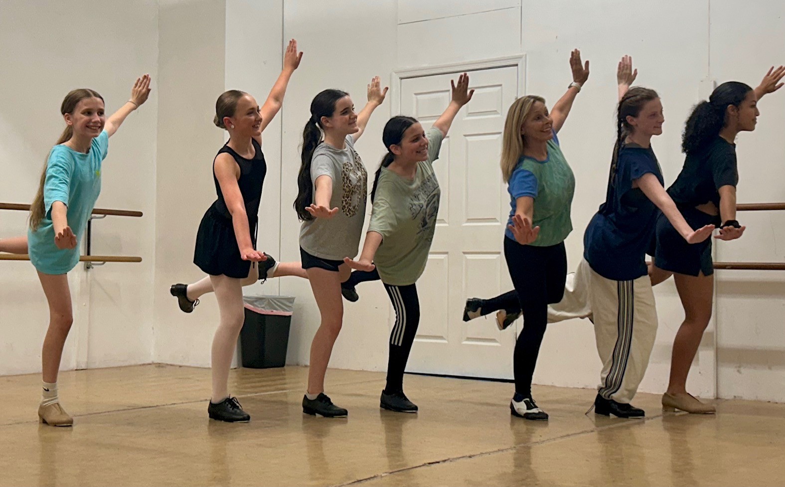 Seven people standing in a line, raising their hands, in a dance studio with wooden floor and white walls.