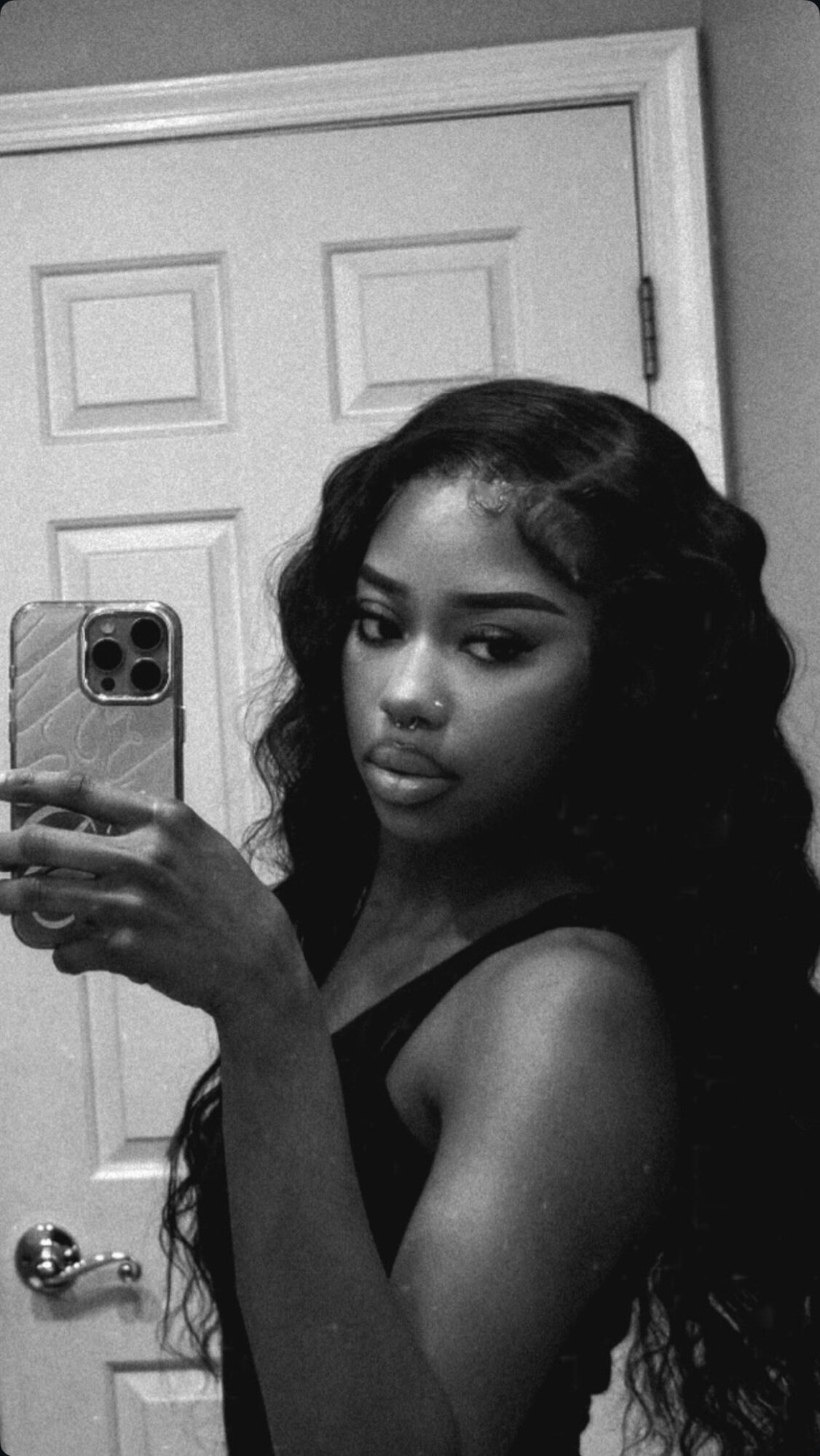 Young woman with long wavy hair taking a mirror selfie in black and white, wearing a sleeveless top.
