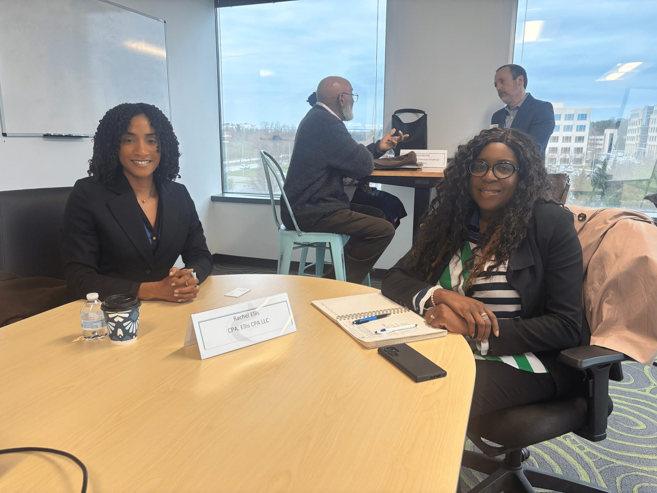 Two women sit at a conference table in a modern office with large windows, smiling at the camera.